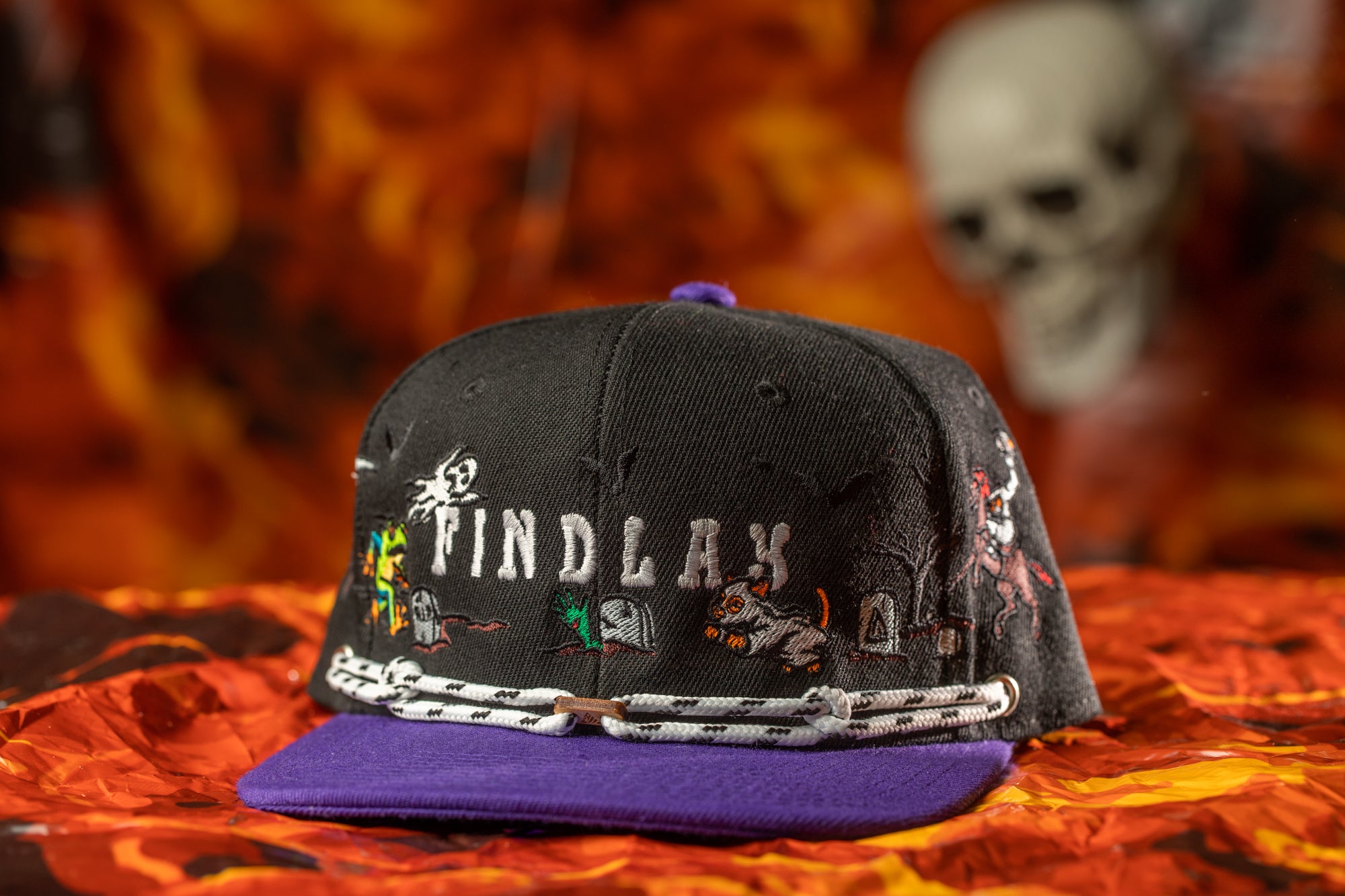 A black baseball cap with a purple brim, featuring various cartoon-like characters and designs, is displayed against a fiery background.