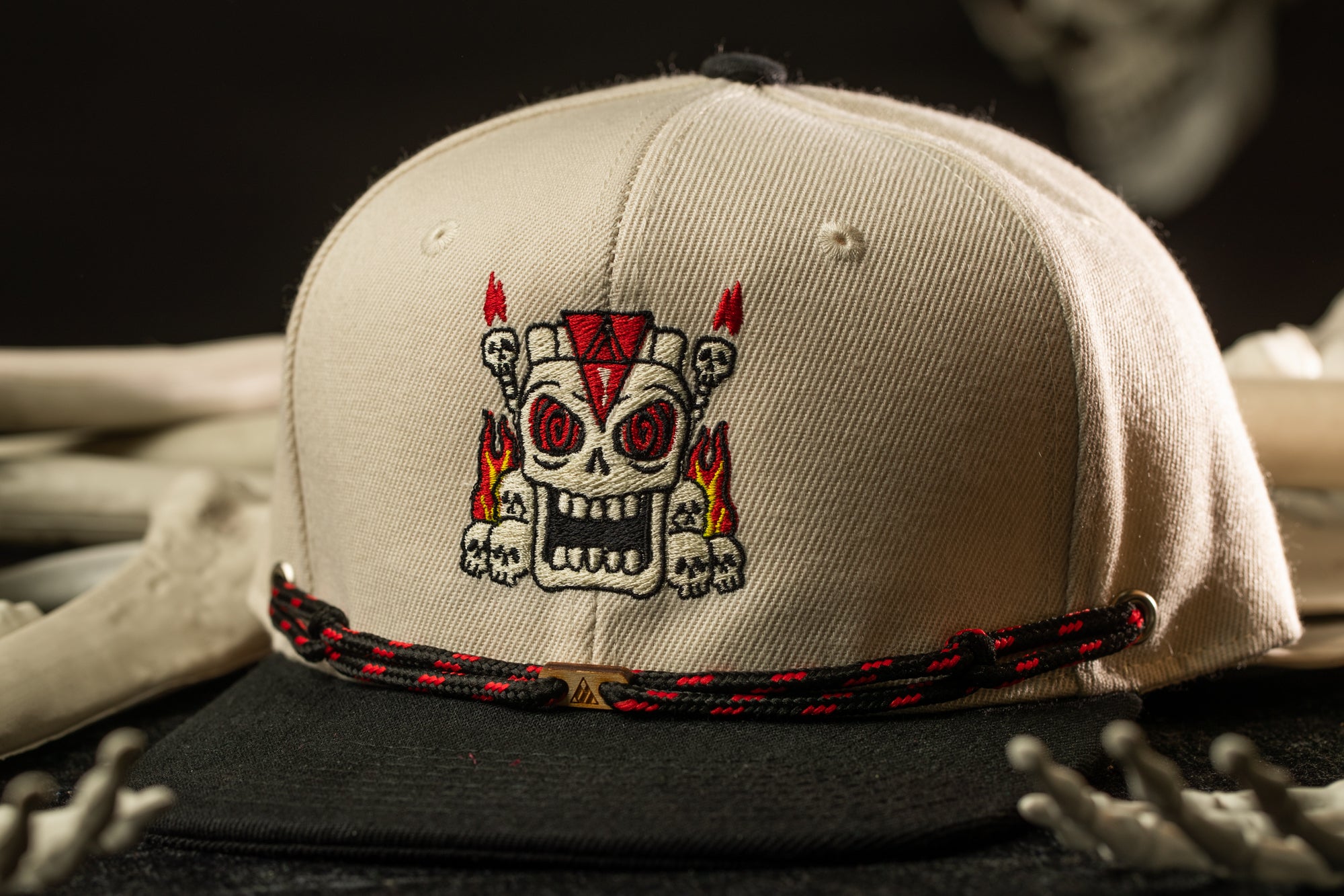 A beige baseball cap with a skull design and a red and black braided strap.