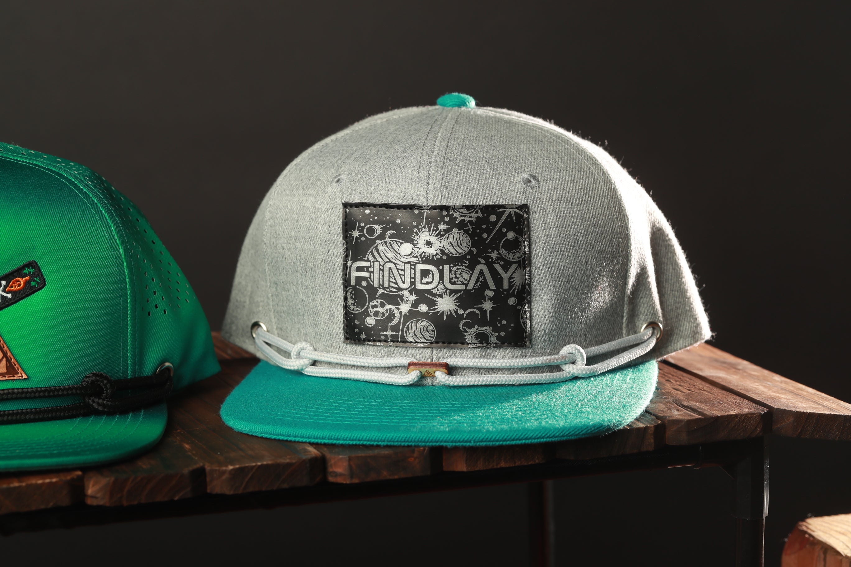A gray baseball cap with a teal brim and a black and white graphic on the front, sitting on a wooden surface.