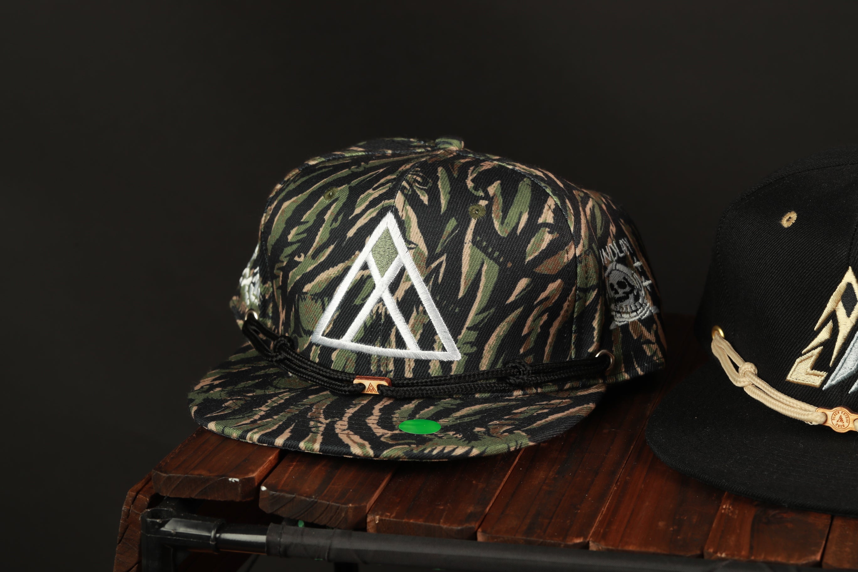 two baseball caps on a wooden surface, one with a camouflage pattern and the other with a black and gold design.