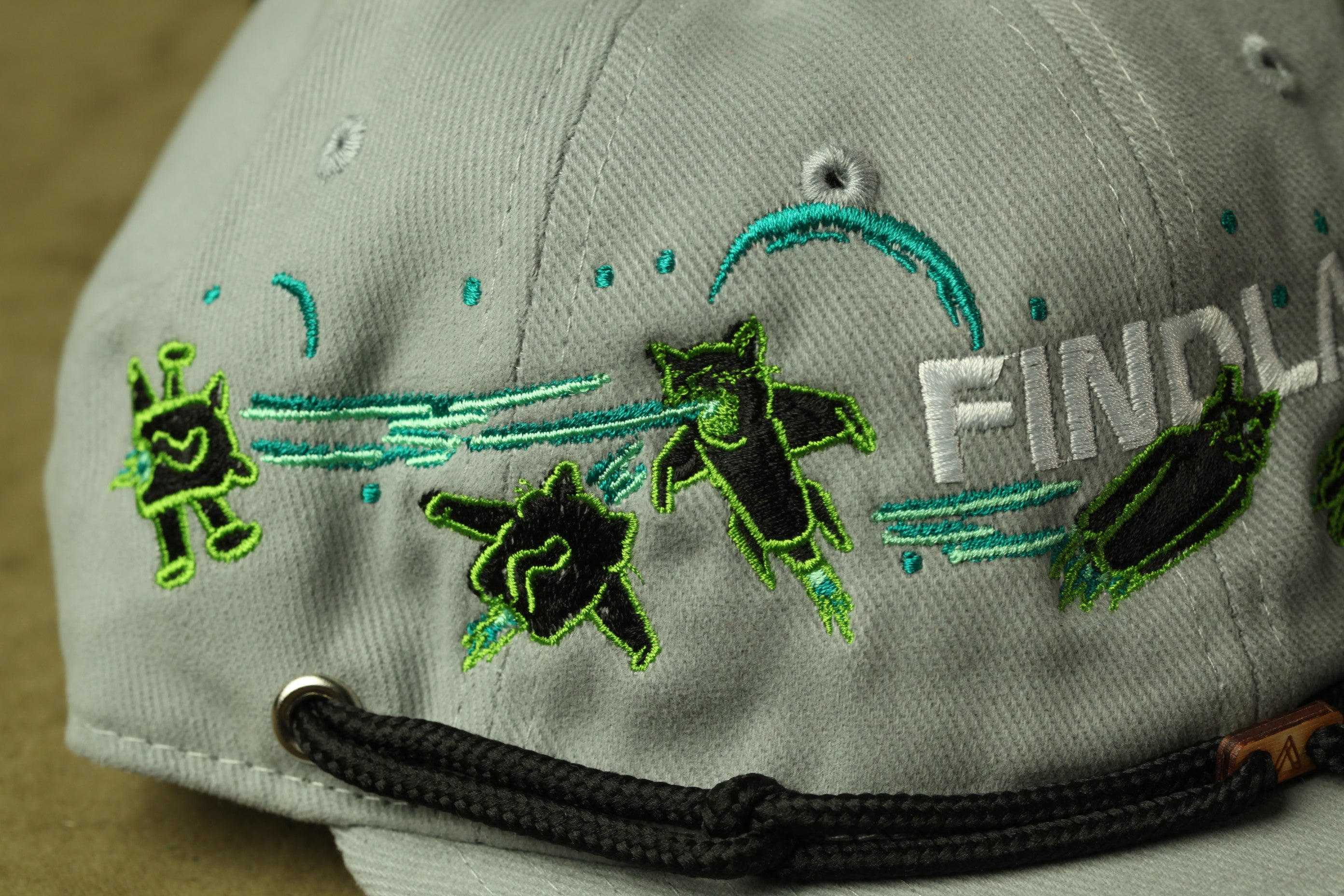 A gray baseball cap with green and black embroidered designs, including a character and the word "FIND".