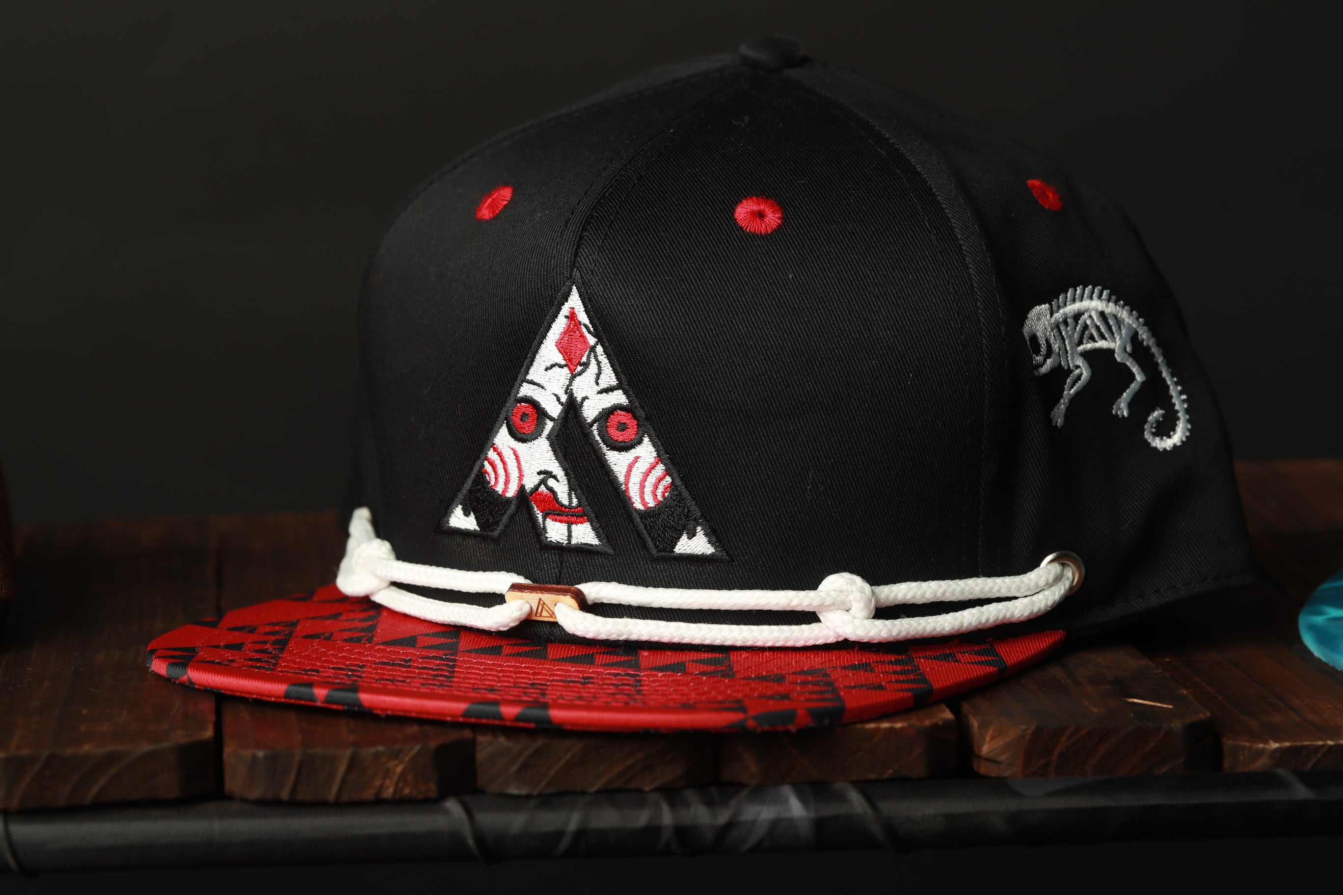 A black baseball cap with a red and white design on the front, featuring a stylized letter 'A' and a dinosaur-like figure.