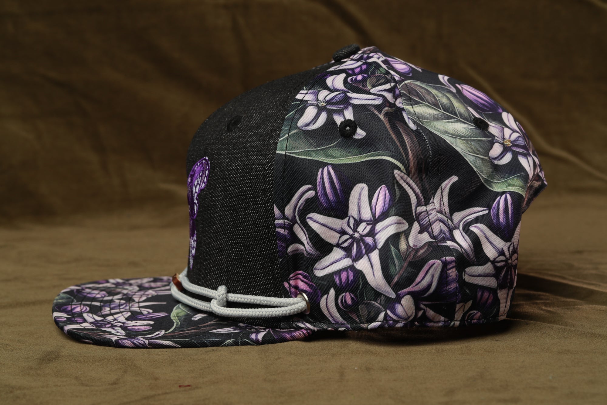 A black and purple floral baseball cap with a white string, resting on a brown surface.