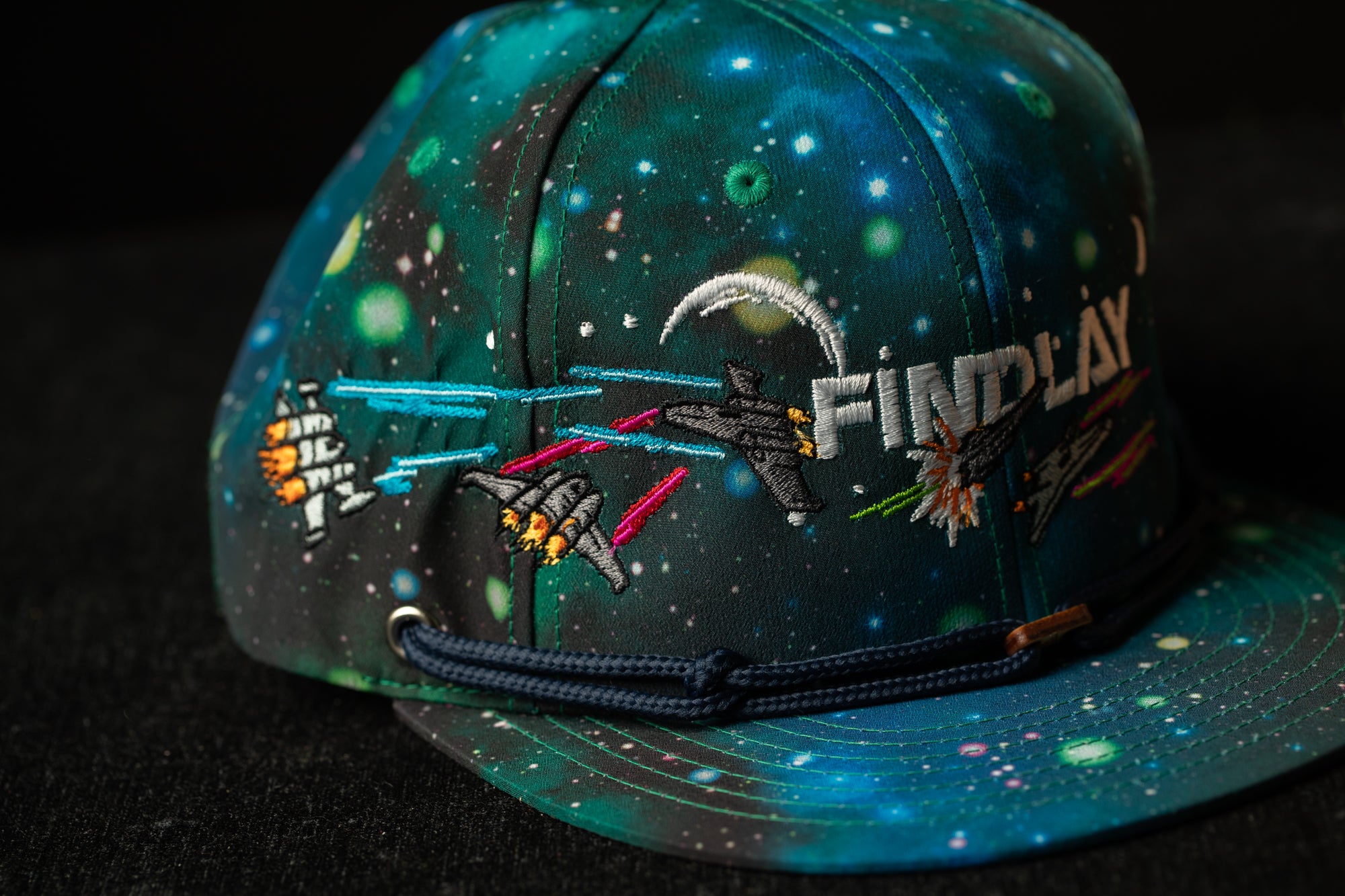 A colorful baseball cap with a space-themed design, featuring various celestial elements such as stars, planets, and a rocket ship.