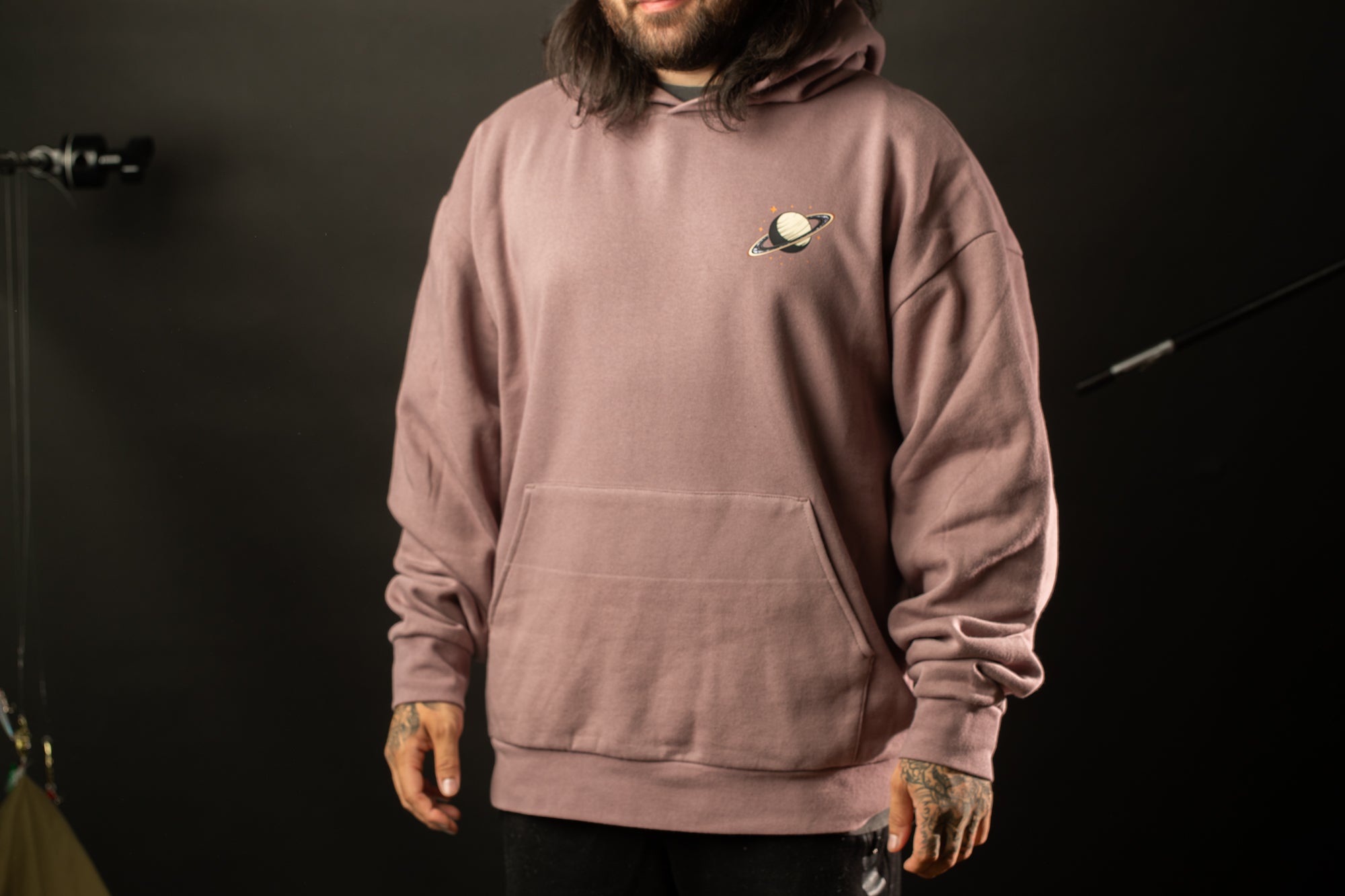 A person wearing a pink hoodie with a planet design on the front stands against a dark background.