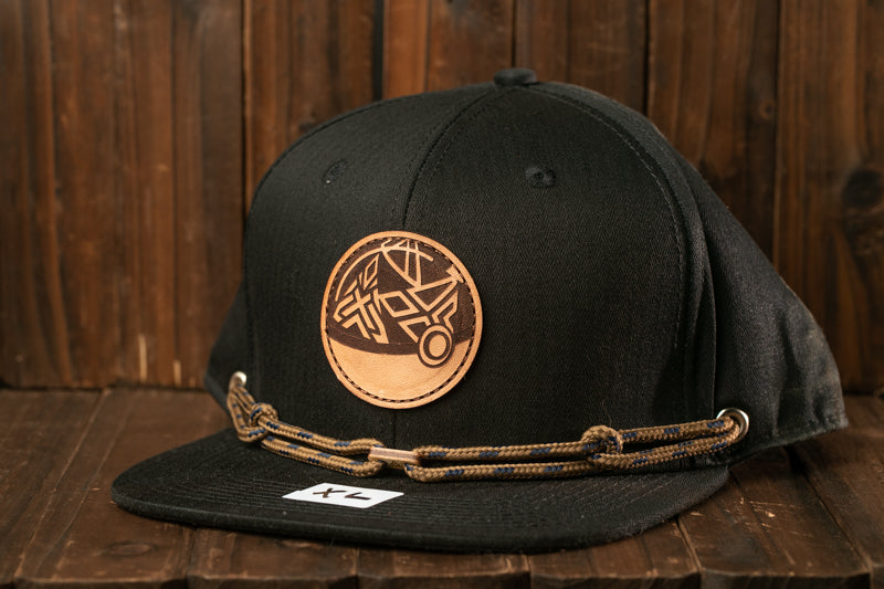 A black baseball cap with a circular logo and a chain strap is displayed on a wooden surface.
