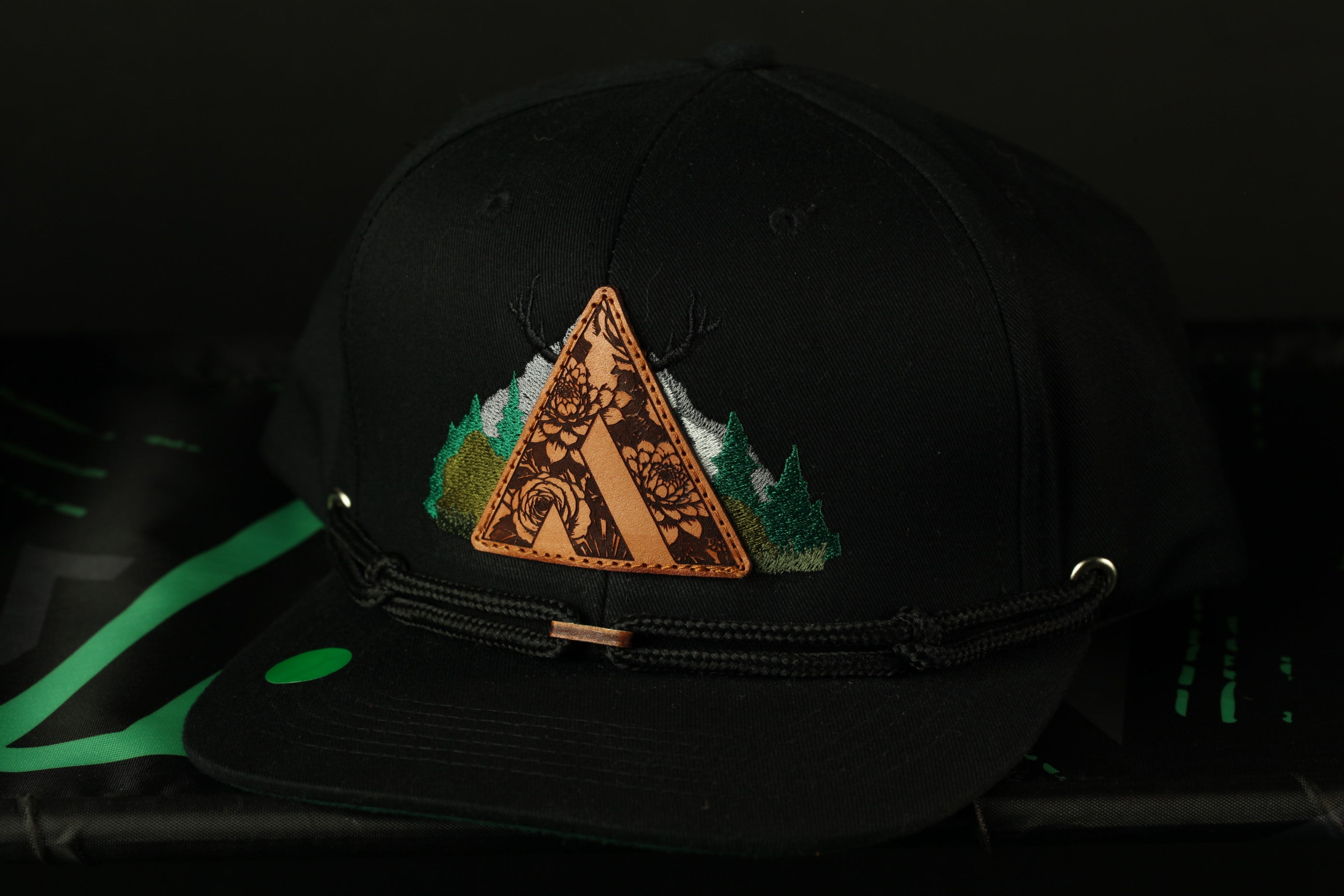 A black baseball cap with a decorative triangle design on the front, featuring a floral pattern and a mountain landscape.