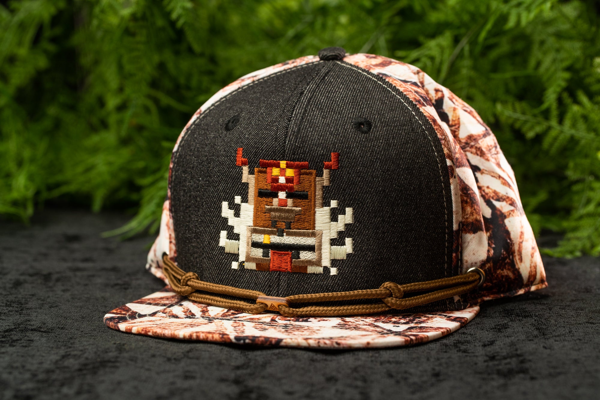 A black baseball cap with a pixelated design on the front, placed on a surface with greenery in the background.