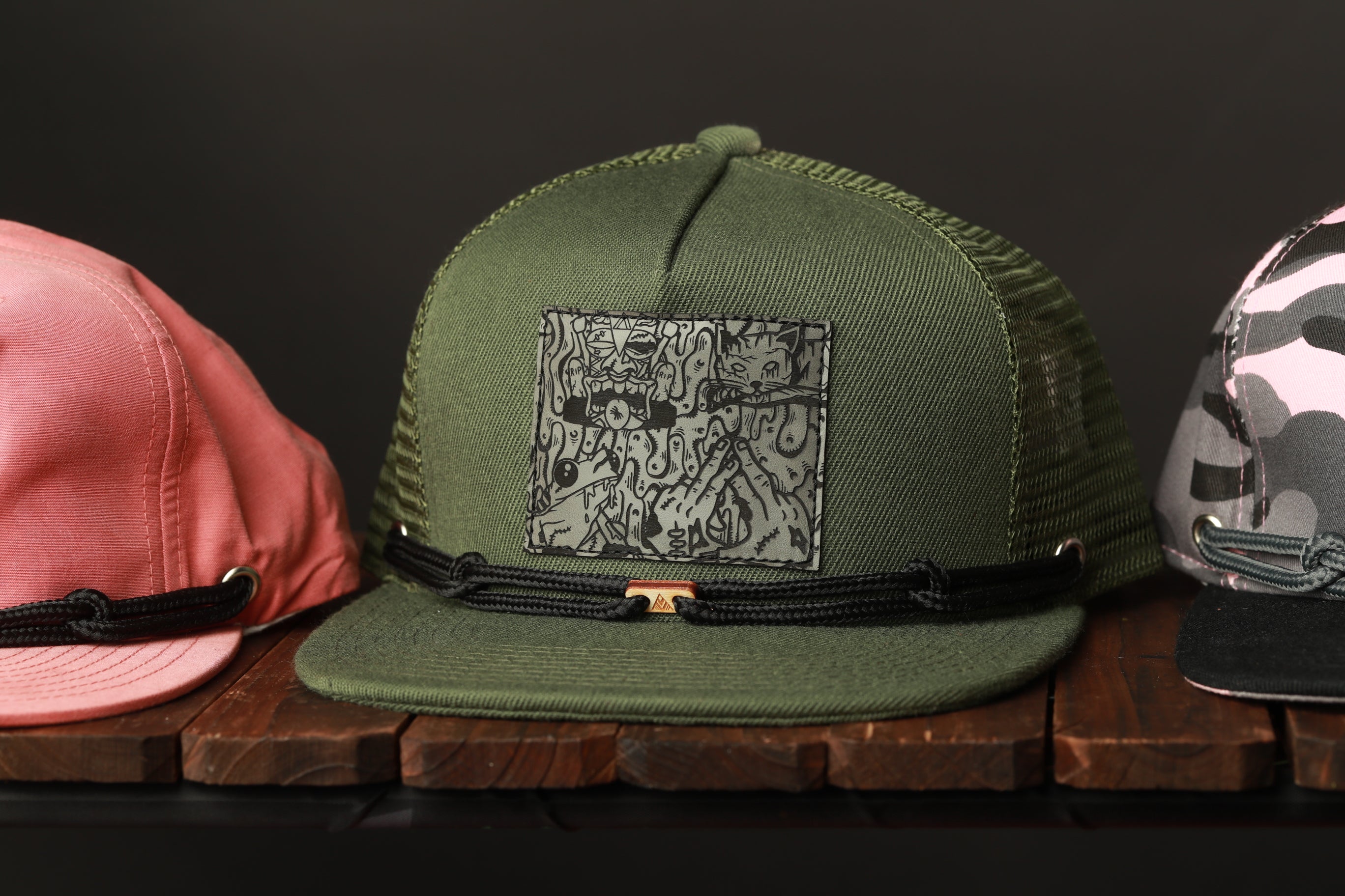 Three different hats are displayed on a wooden surface, with one green hat featuring a graphic design and two other hats in the background.