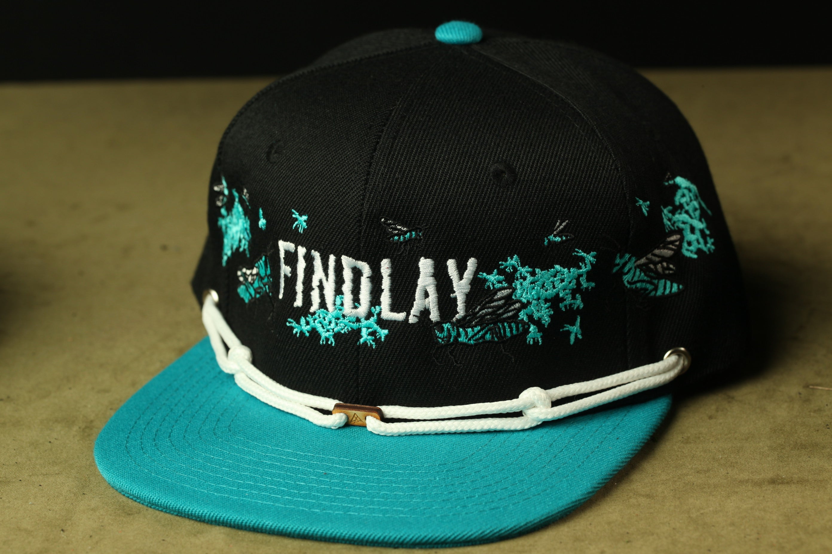 A black baseball cap with teal trim and the word "FINDLAY" embroidered on it.