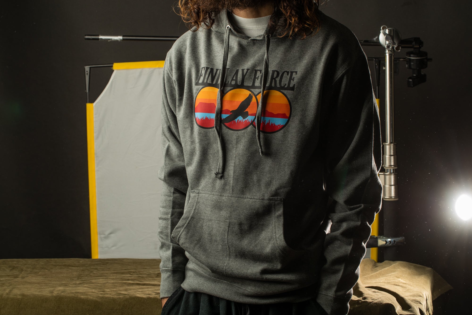 A person wearing a grey hoodie with a graphic design on it, standing in a dark room with a camera and other equipment in the background.