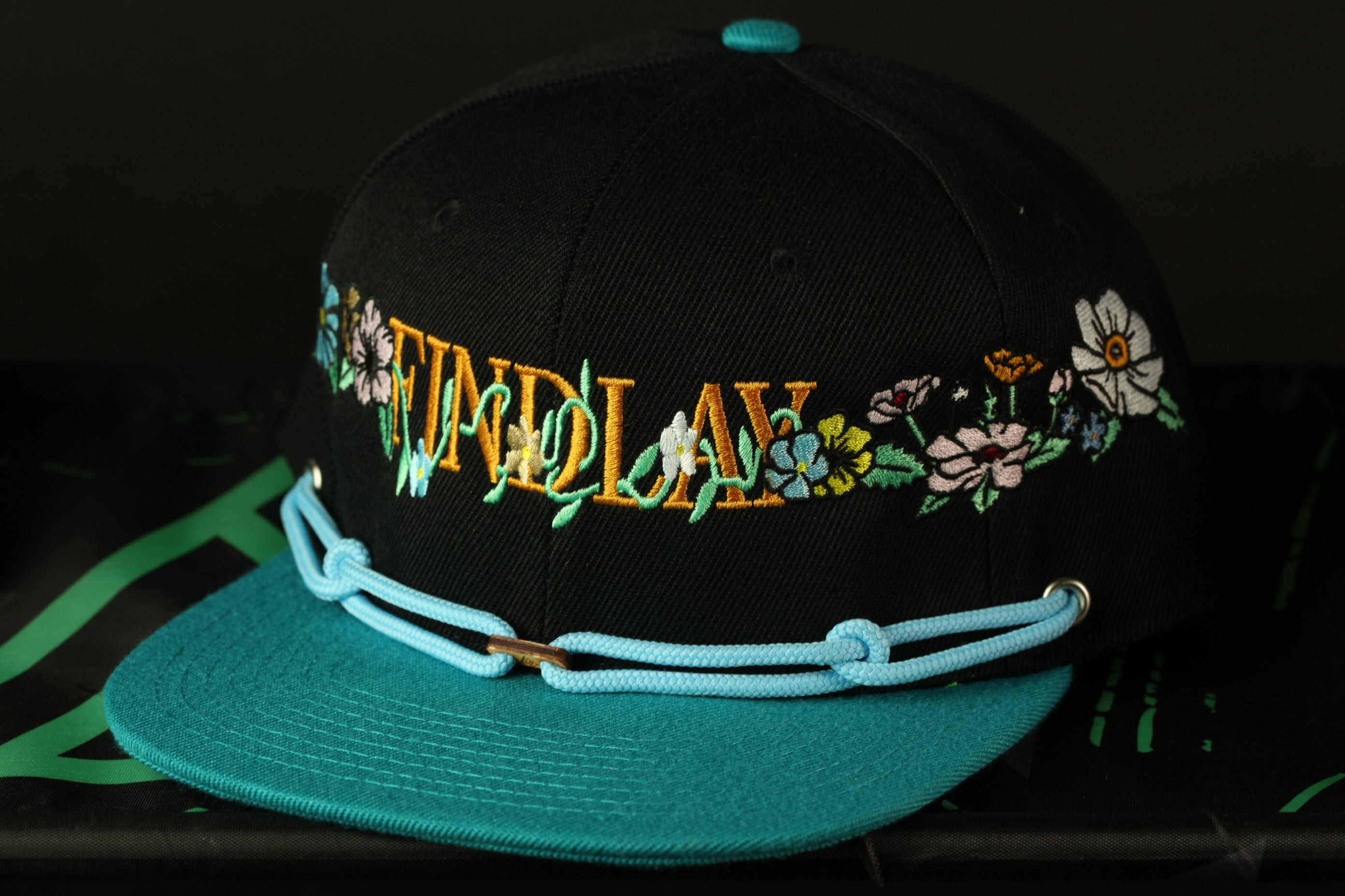 A black baseball cap with a teal brim and floral embroidered text on the front.