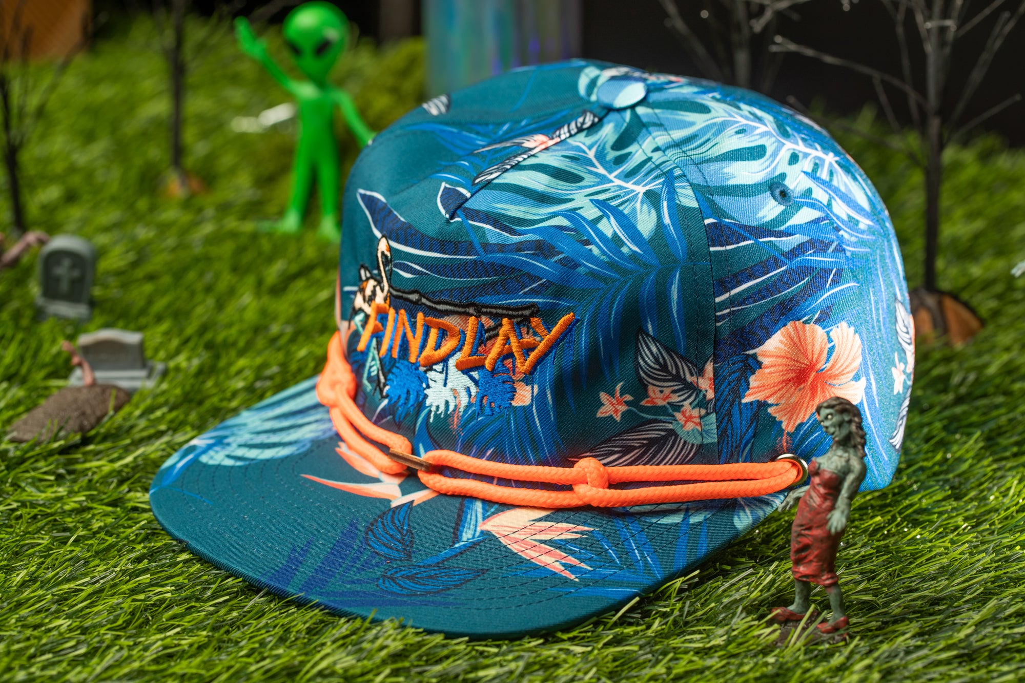 A colorful tropical-themed baseball cap with the word "INDLAY" printed on it, placed on a grassy surface with small figurines and a green alien figure in the background.