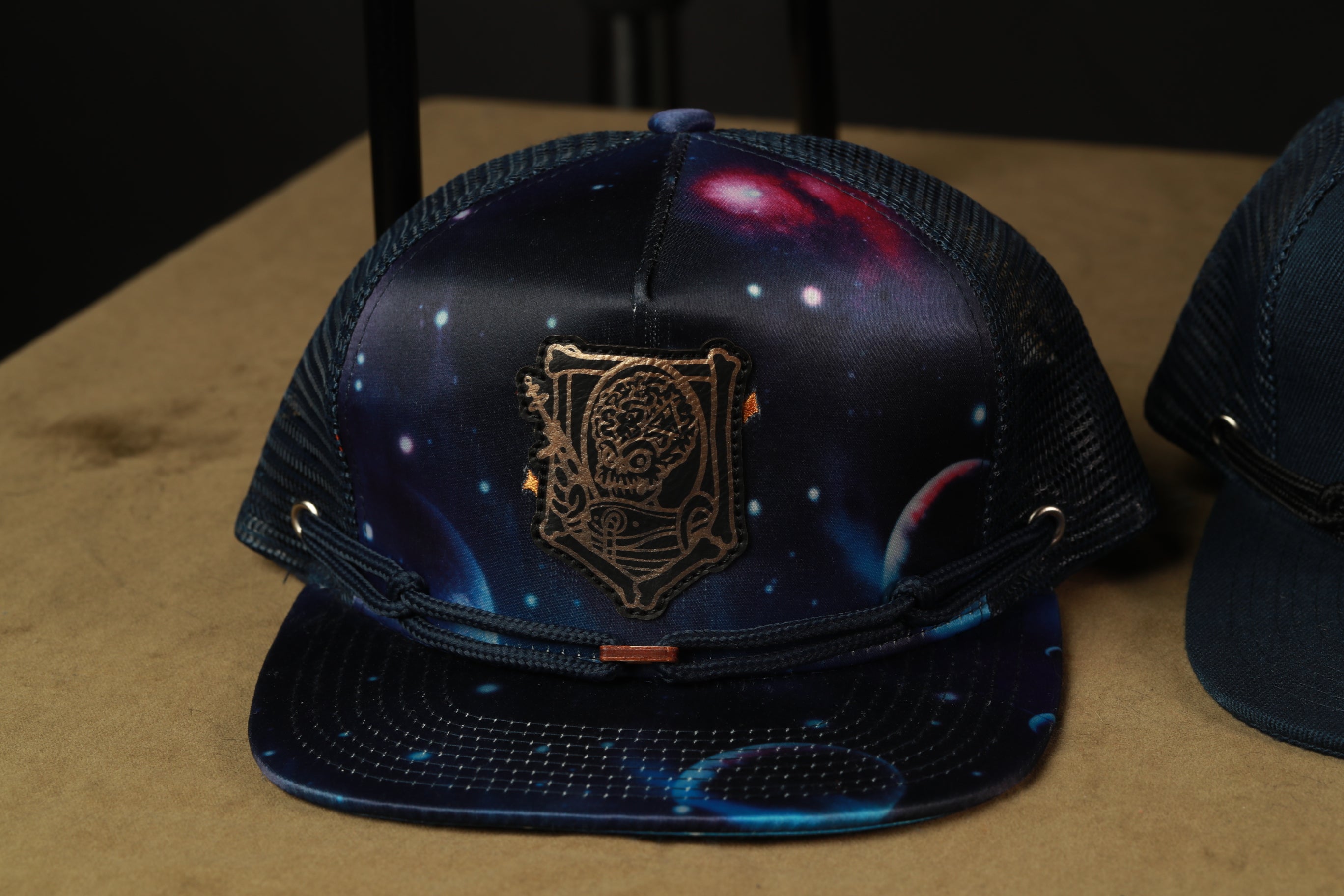 A black baseball cap with a space-themed design, featuring a central emblem and a starry night sky pattern.