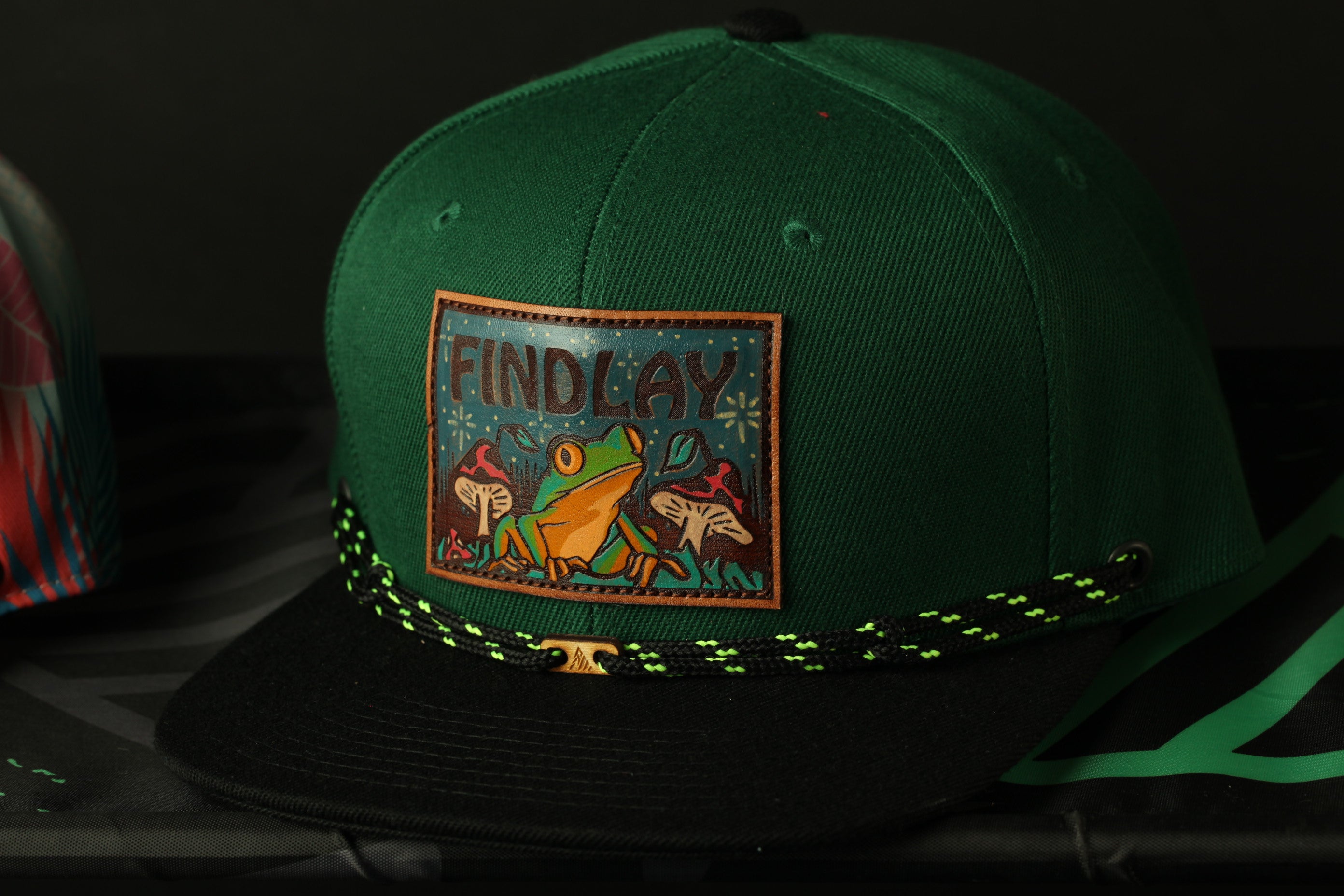 A green baseball cap with a patch featuring a frog and mushrooms, along with the text "FINDLAY".