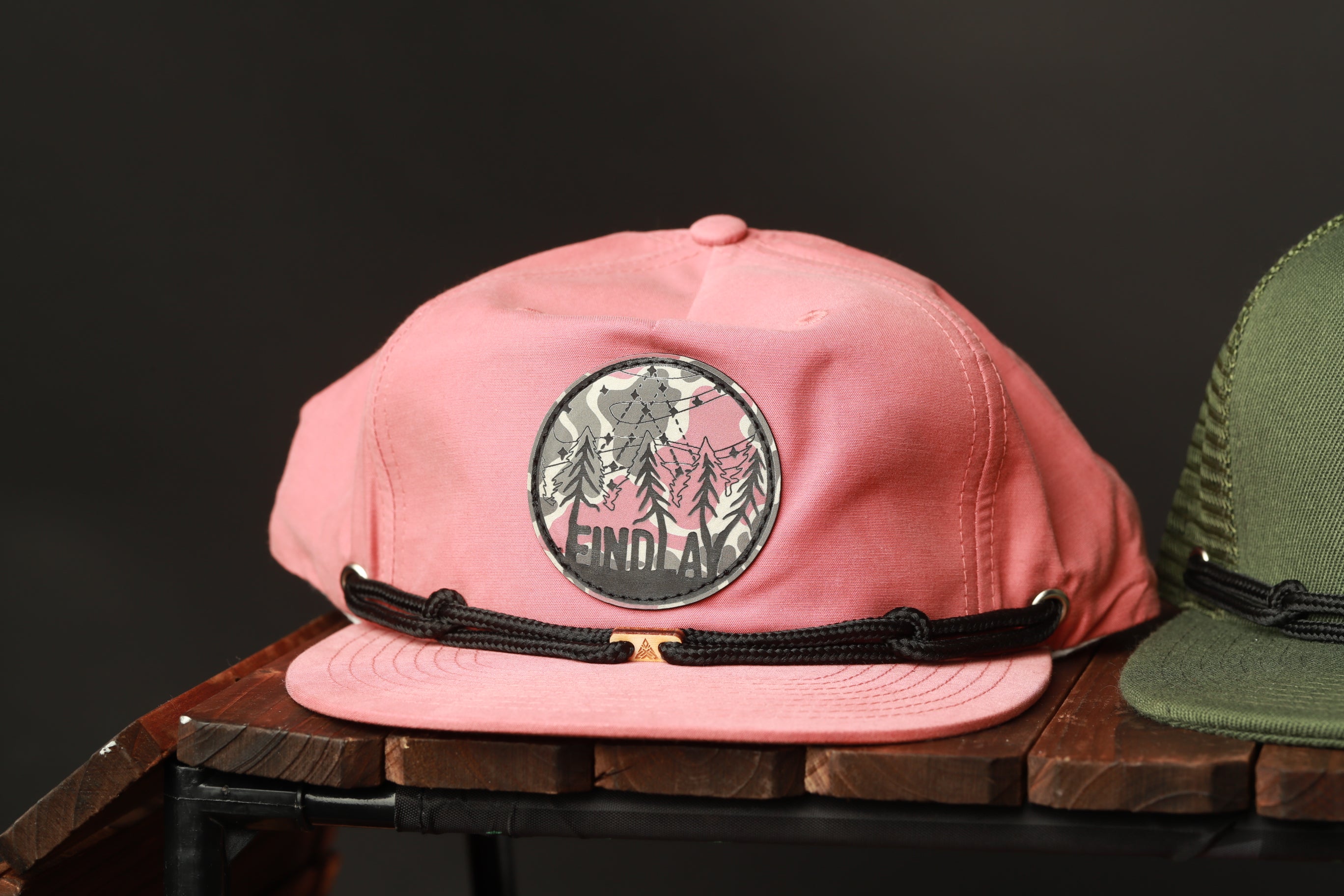A pink baseball cap with a circular patch on the front displaying the word "FINDLAK" is placed on a wooden surface.