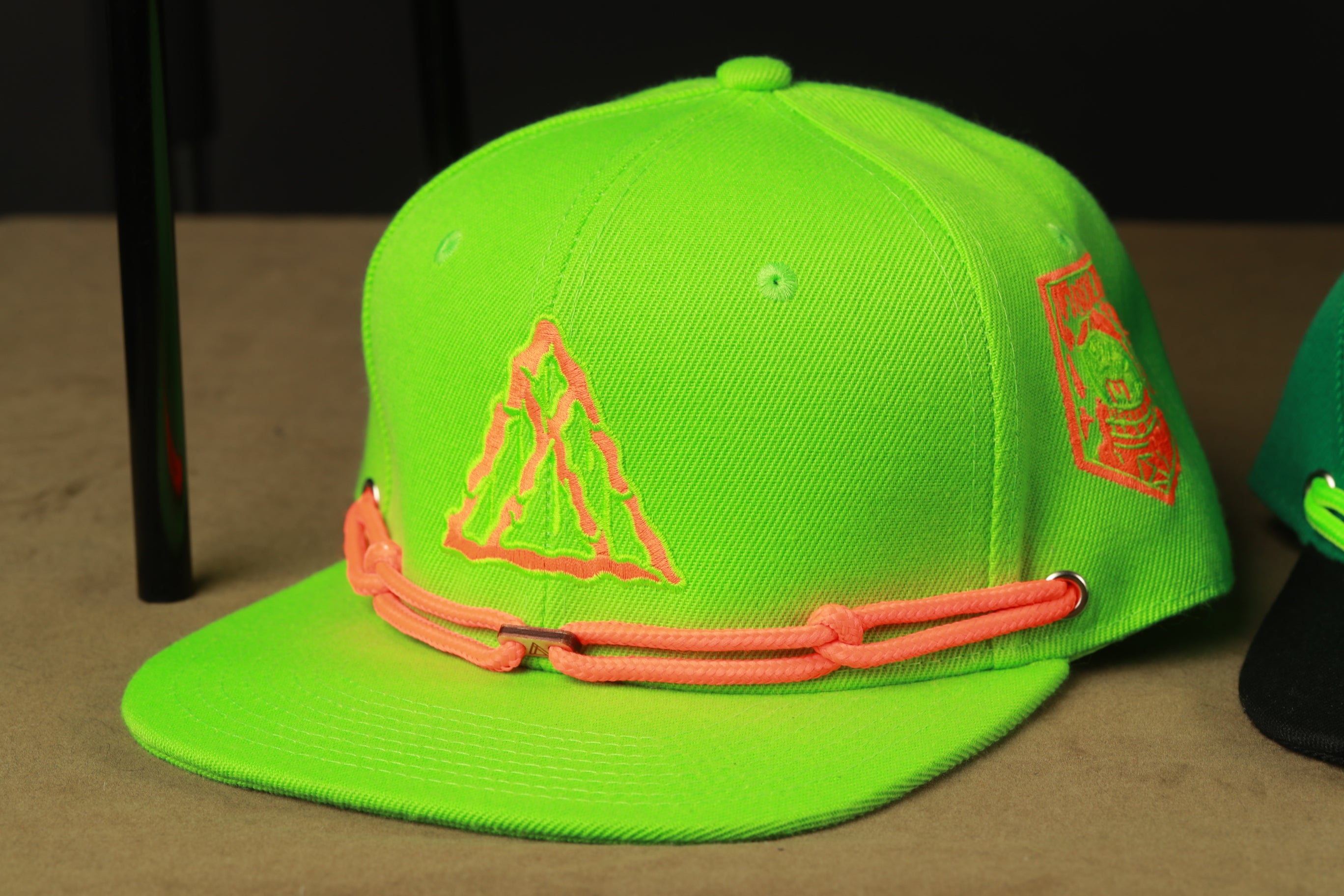 A neon green baseball cap with a triangle logo and orange braided straps is displayed on a table.