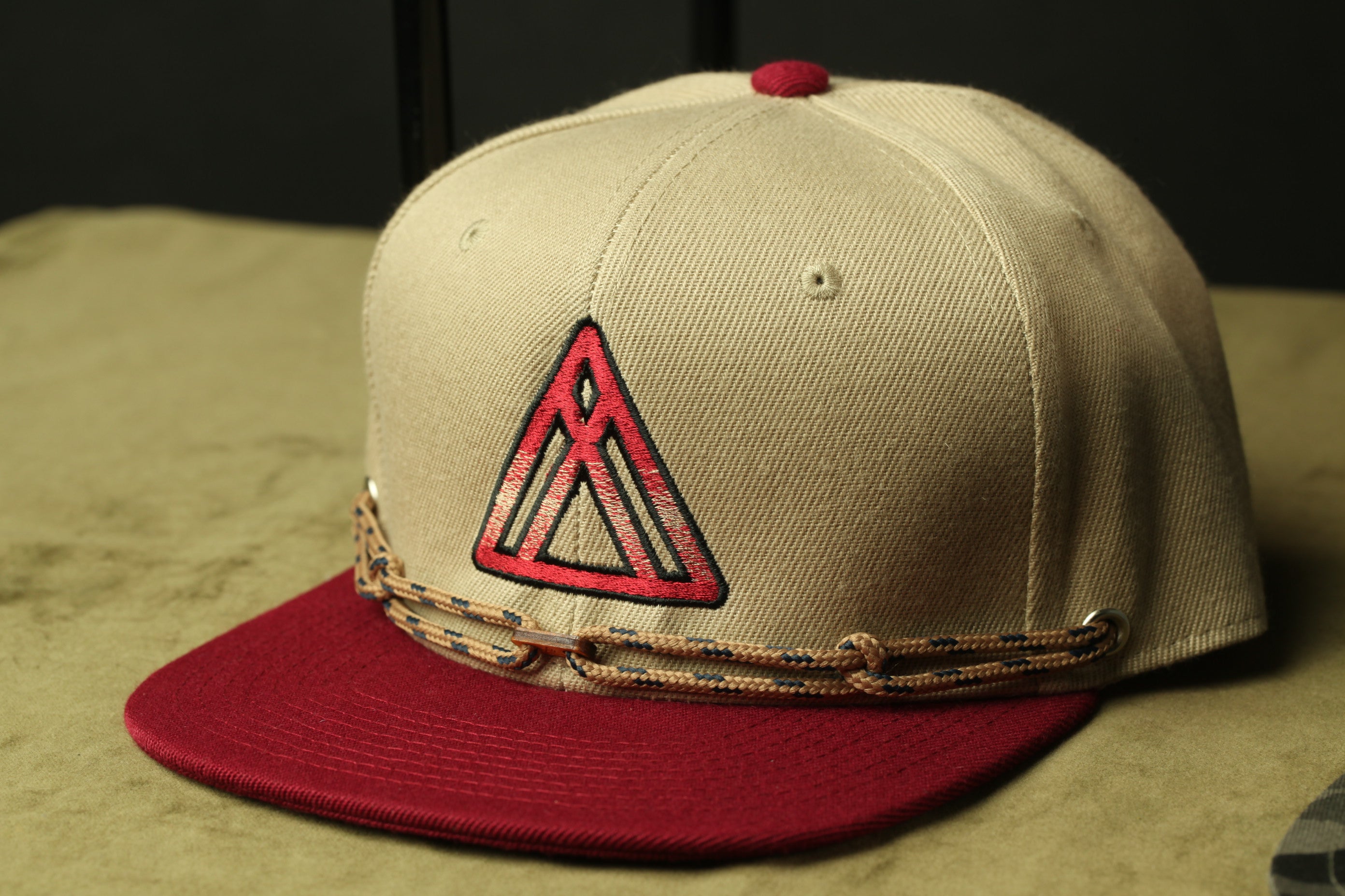 A beige baseball cap with a red triangle logo and a red and maroon woven strap.
