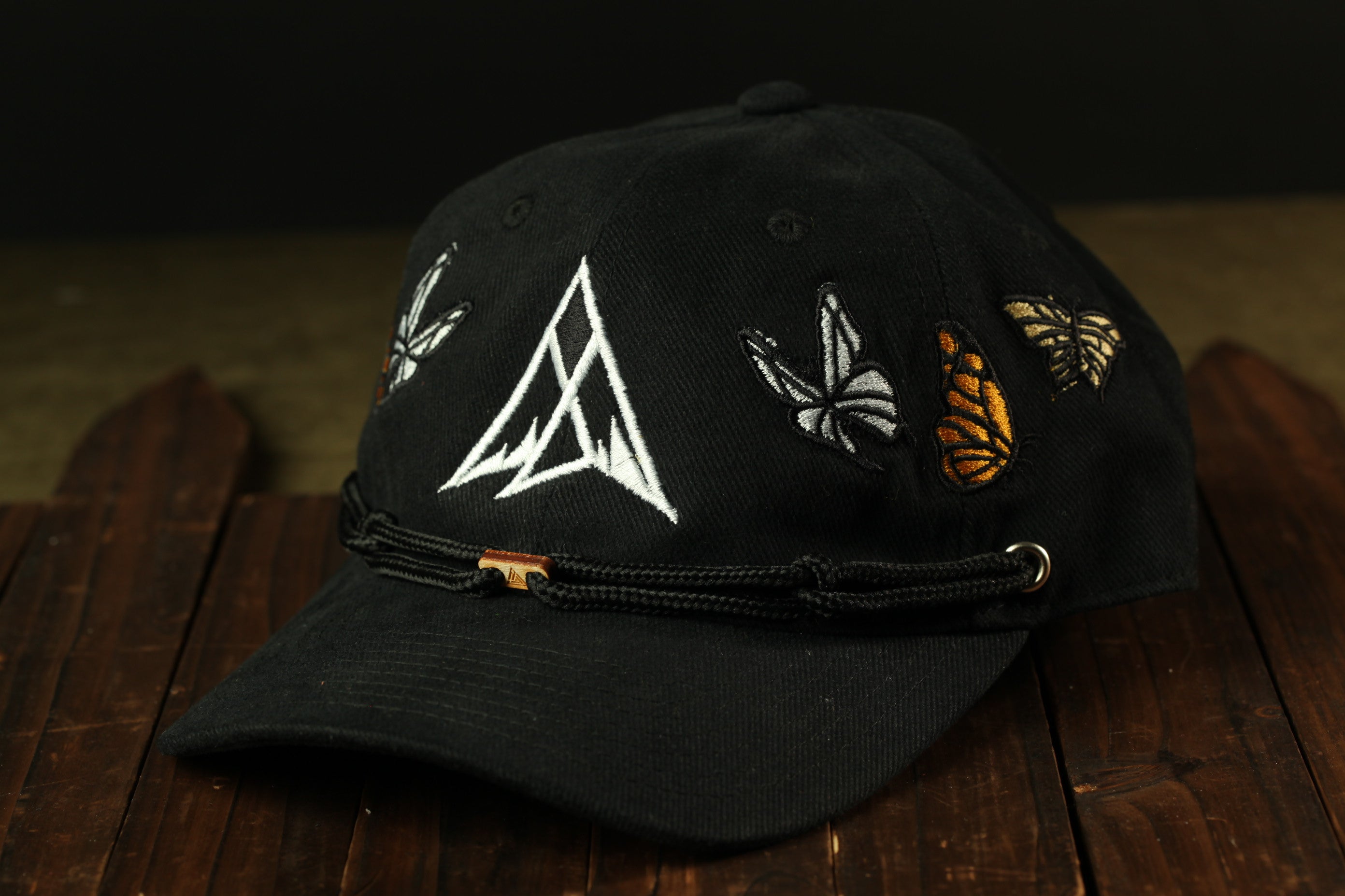 A black baseball cap with a white triangle design and butterflies embroidered on it, resting on a wooden surface.