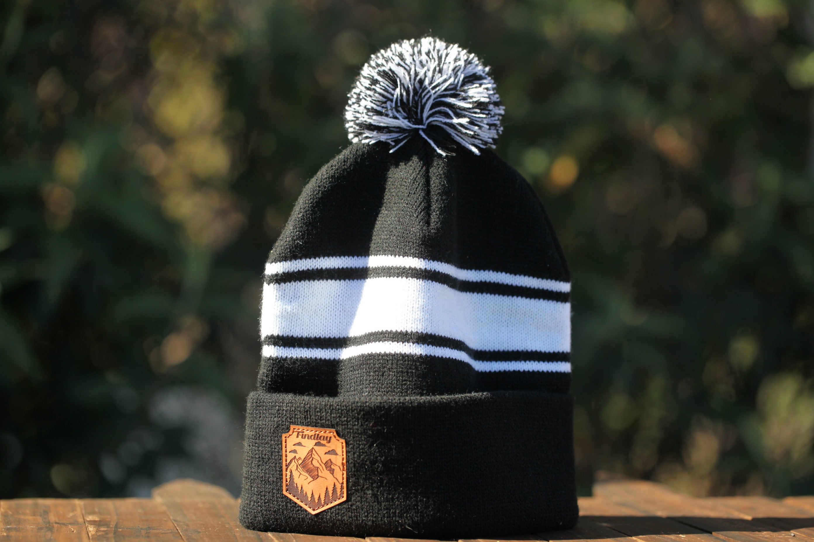 A black and white striped beanie with a pom pom on top, placed on a wooden surface against a blurred natural background.