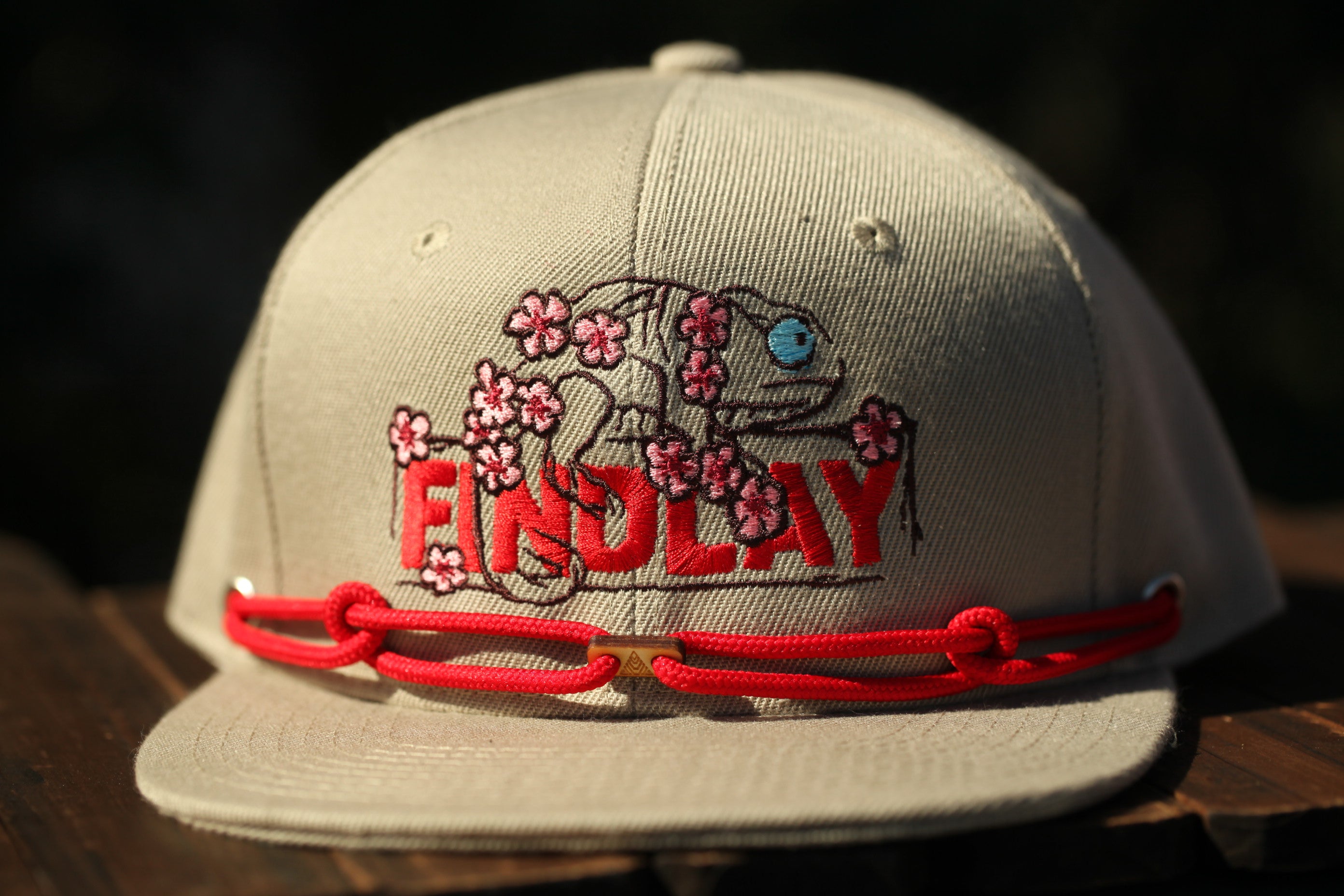 A beige baseball cap with a red string and a small embroidered design on the front, featuring the word "FINDlay" in red letters.