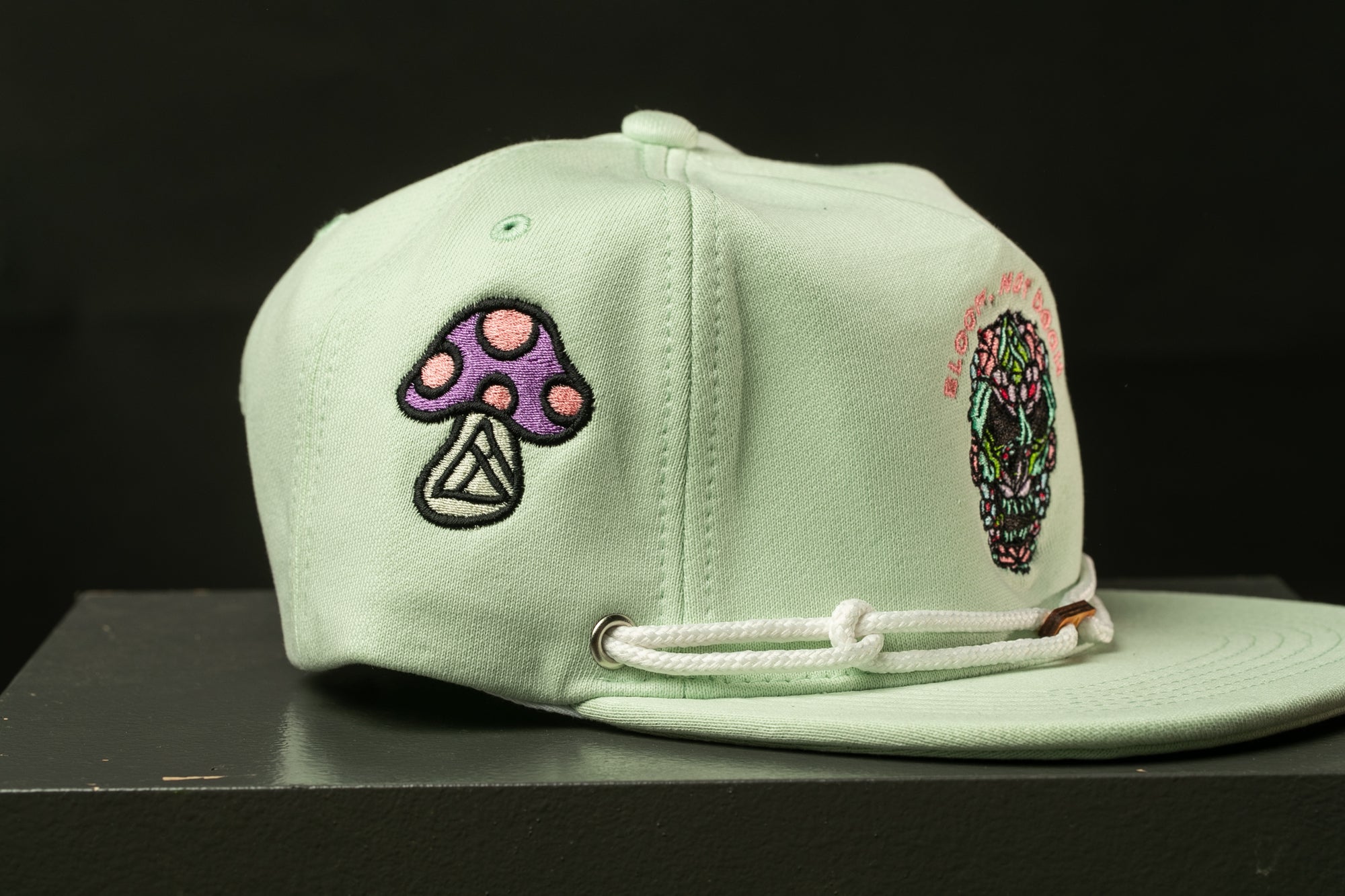 A light green baseball cap with various colorful and intricate embroidered designs, including a mushroom, a skull, and other abstract patterns.