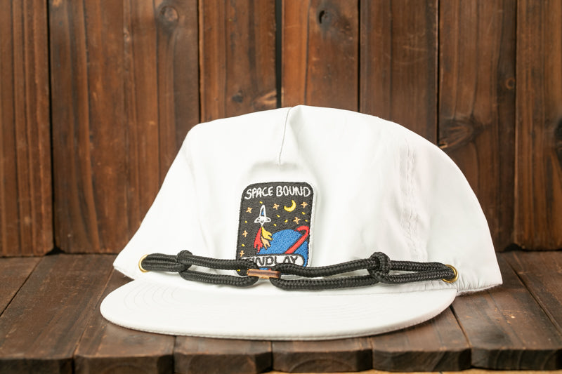 A white baseball cap with a space-themed patch and a black strap is displayed on a wooden surface.