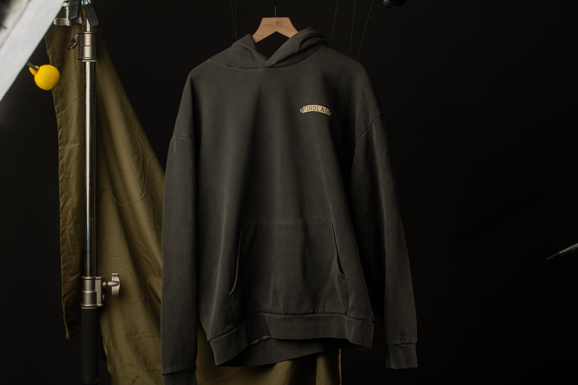 A dark grey hoodie with a small logo on the left side is hanging on a wooden hanger against a black background.