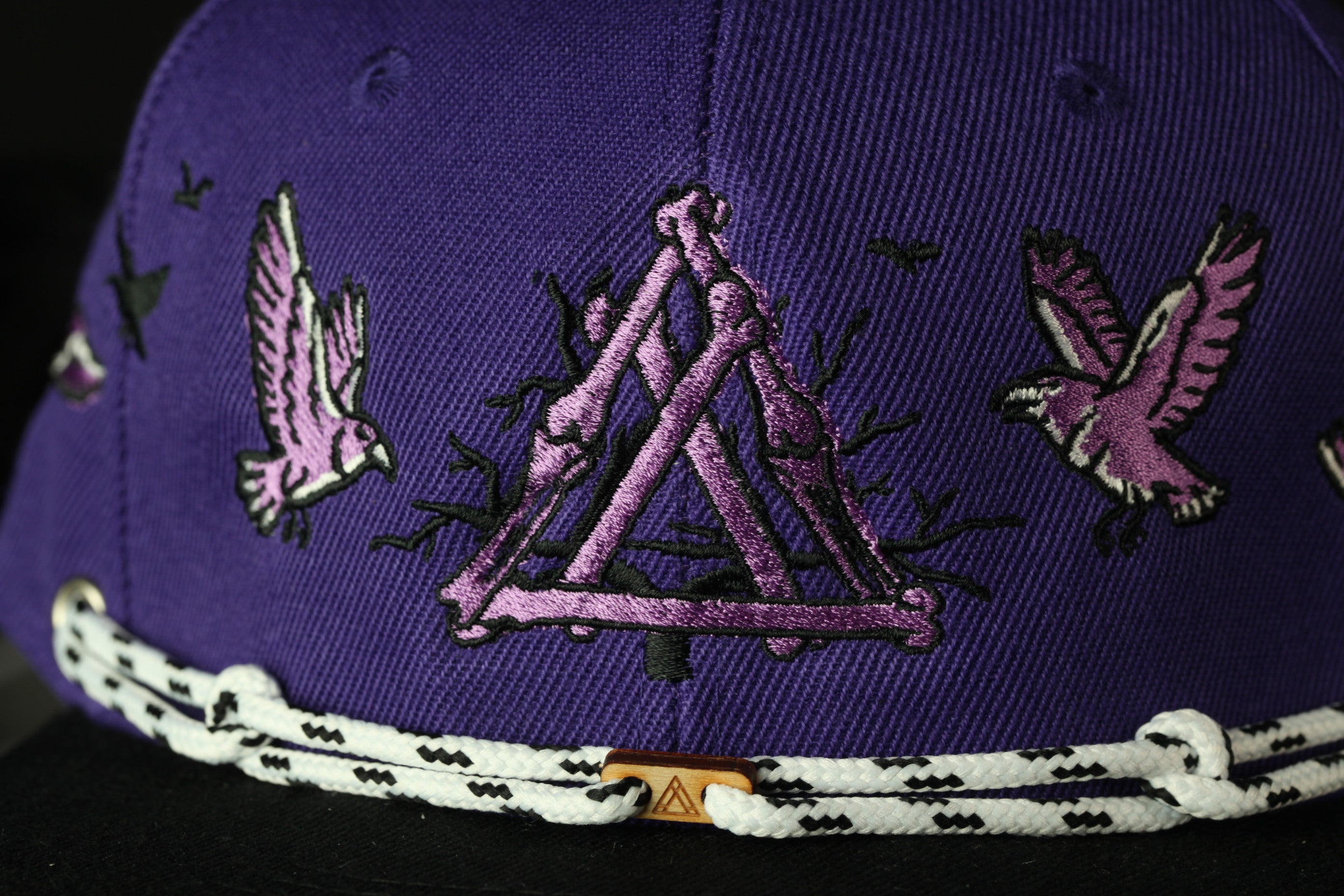 A close-up view of a purple baseball cap with a black and white rope trim, featuring a purple triangle design with a bird motif.
