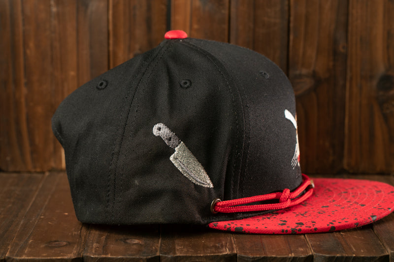 A black baseball cap with a red brim and a knife embroidered on the side, placed on a wooden surface.