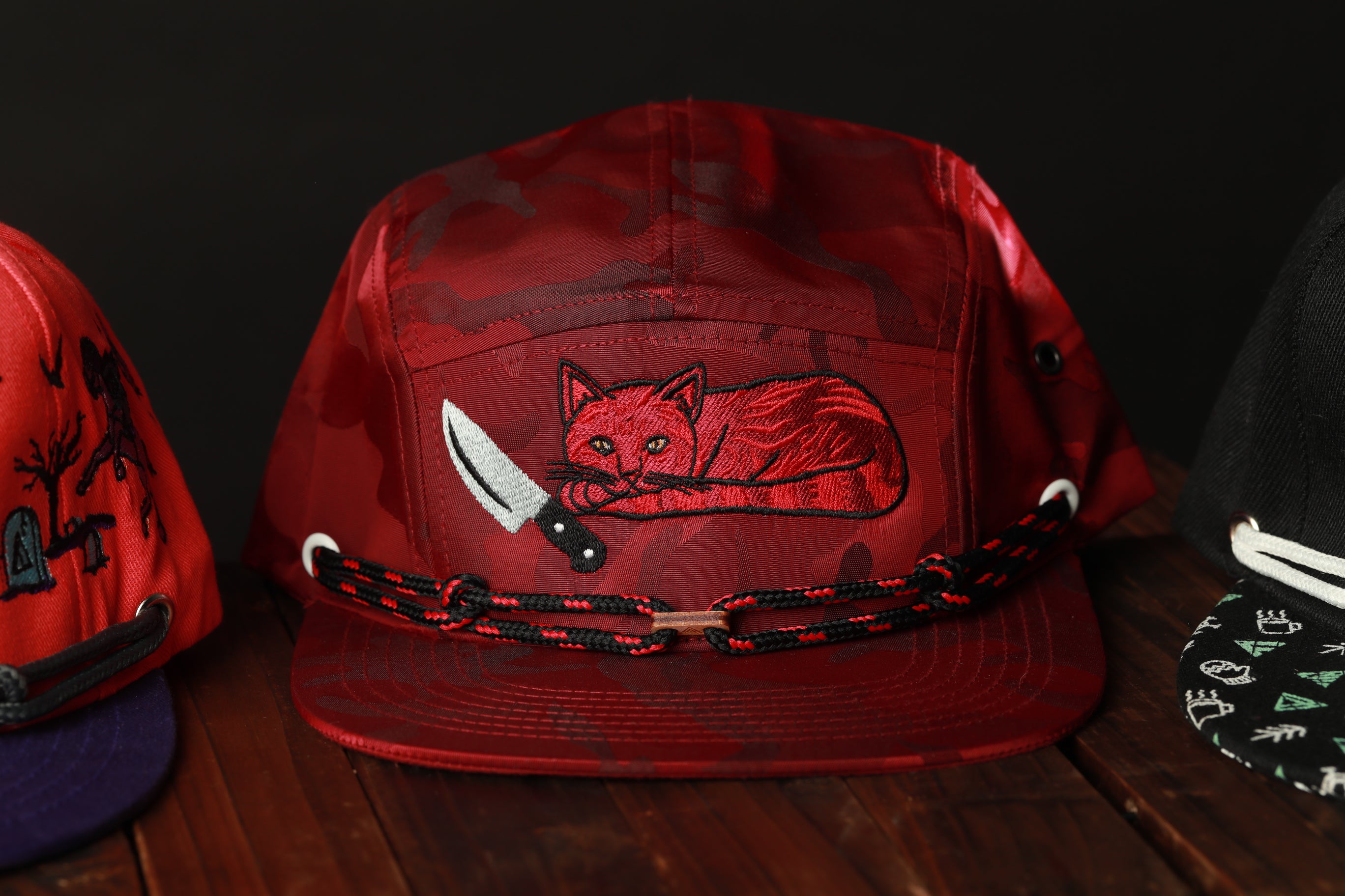 A red baseball cap with a cat embroidered on it, holding a knife, is displayed on a wooden surface.
