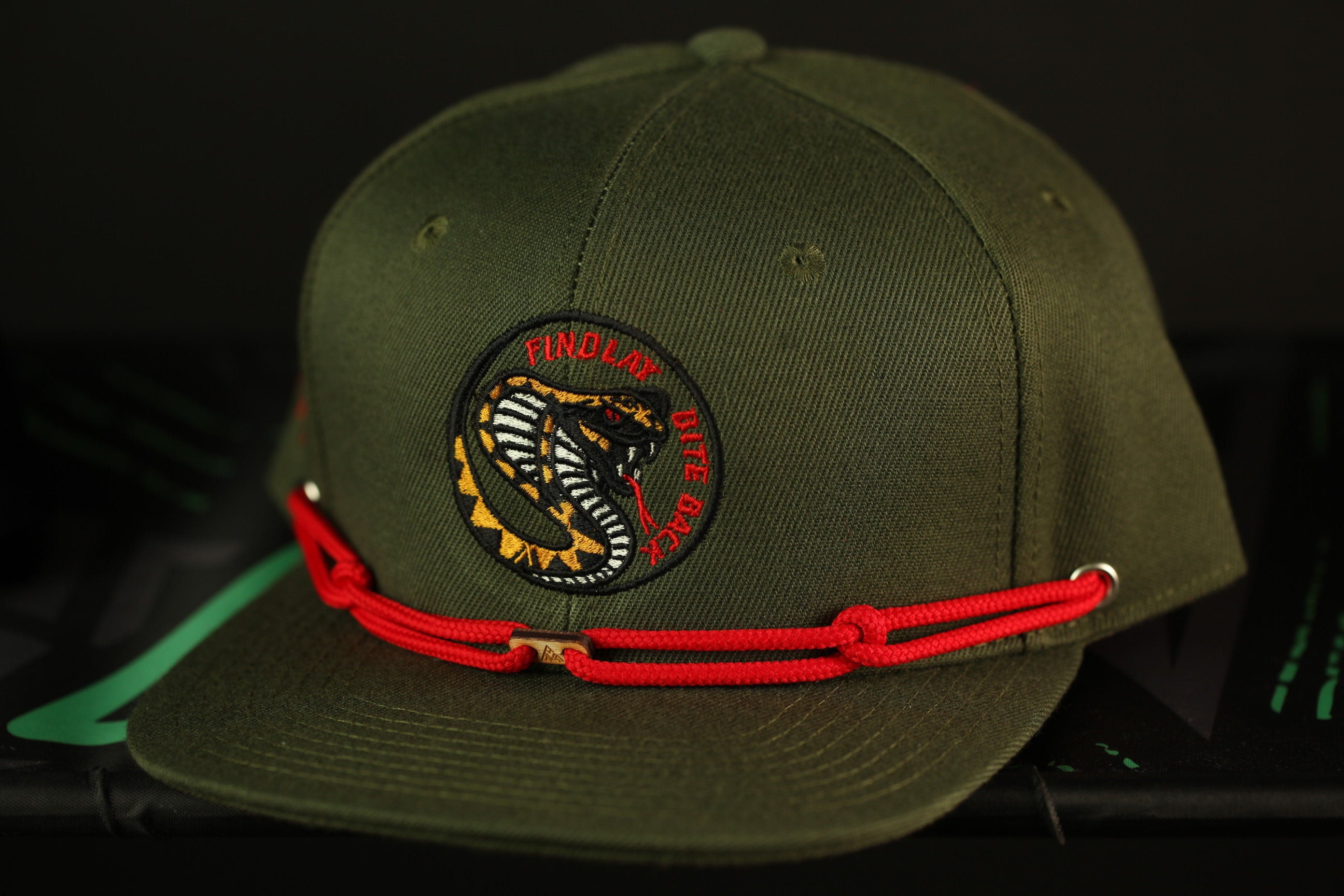A green baseball cap with a red rope and a circular patch on the front, featuring a snake design.