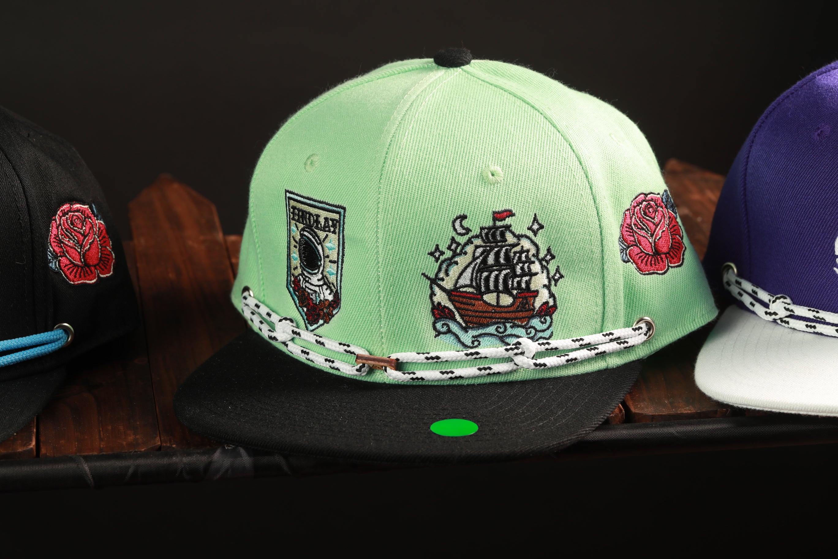 A collection of colorful baseball caps with various designs and patterns, including a green cap with a ship and rose motif, a black cap with a rose and ship design, and a purple cap with a rose and ship design.