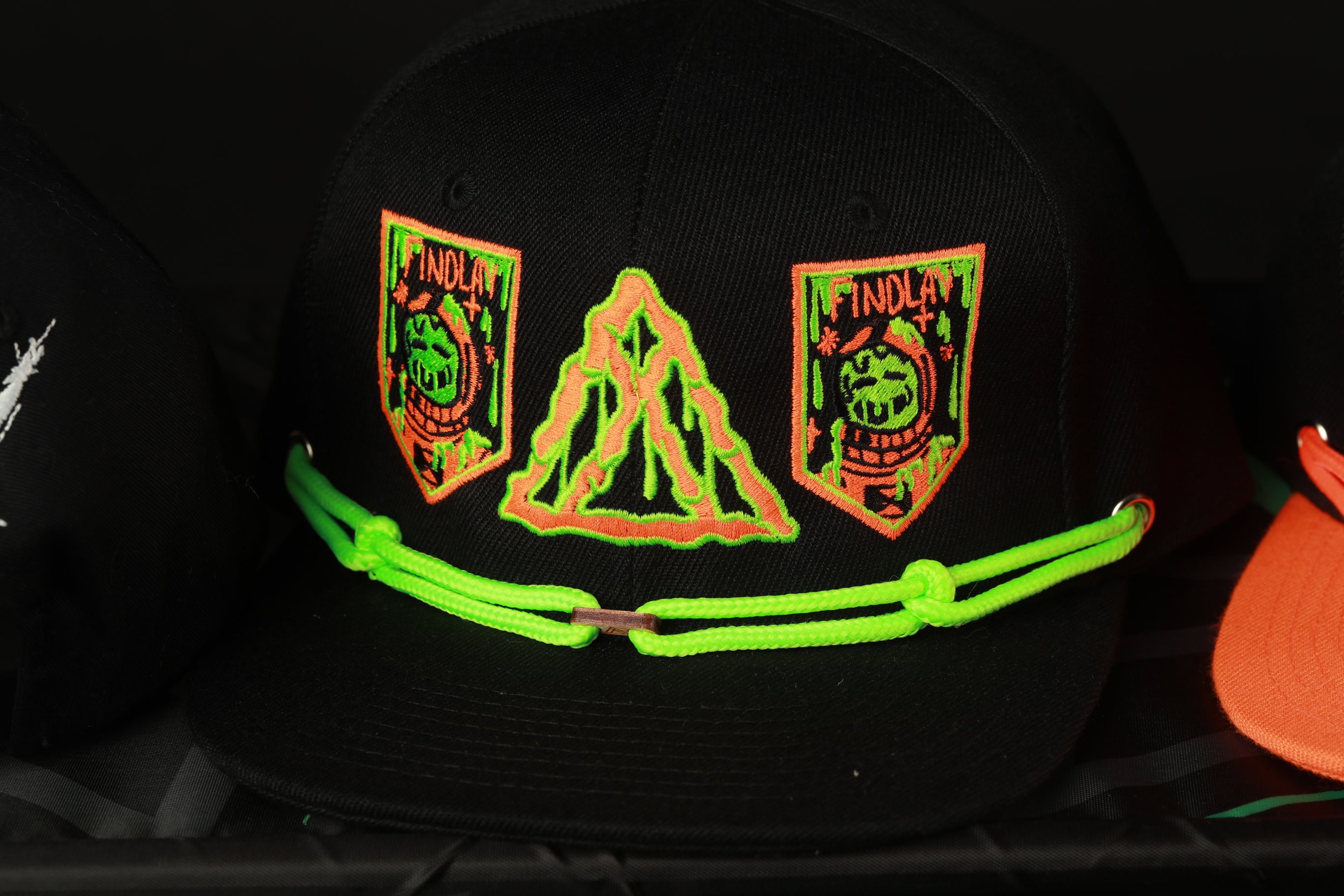 A black baseball cap with neon green and orange patches on it, featuring a triangle design and text.