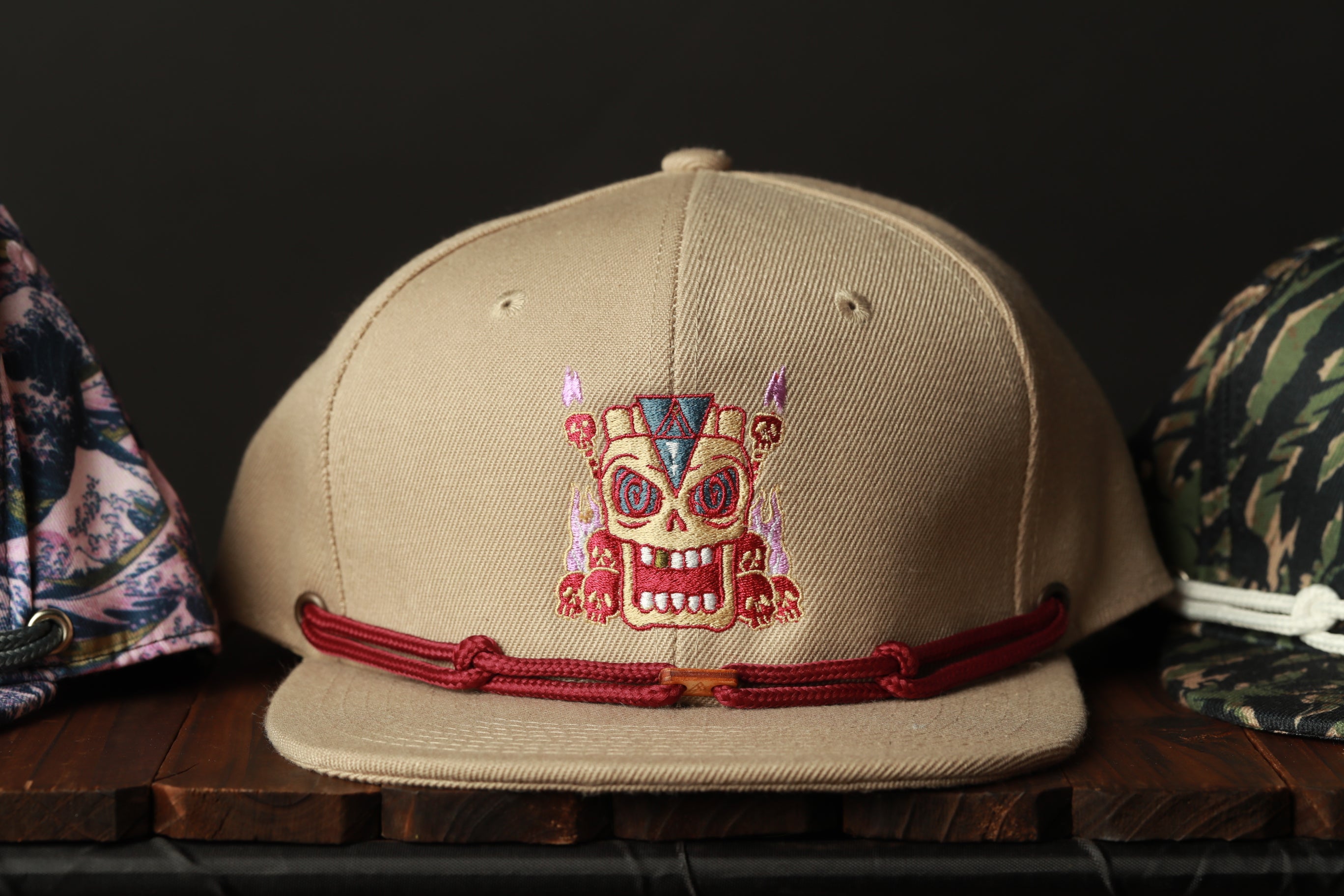 A beige baseball cap with a red string and a skull embroidered on it, displayed on a wooden surface.