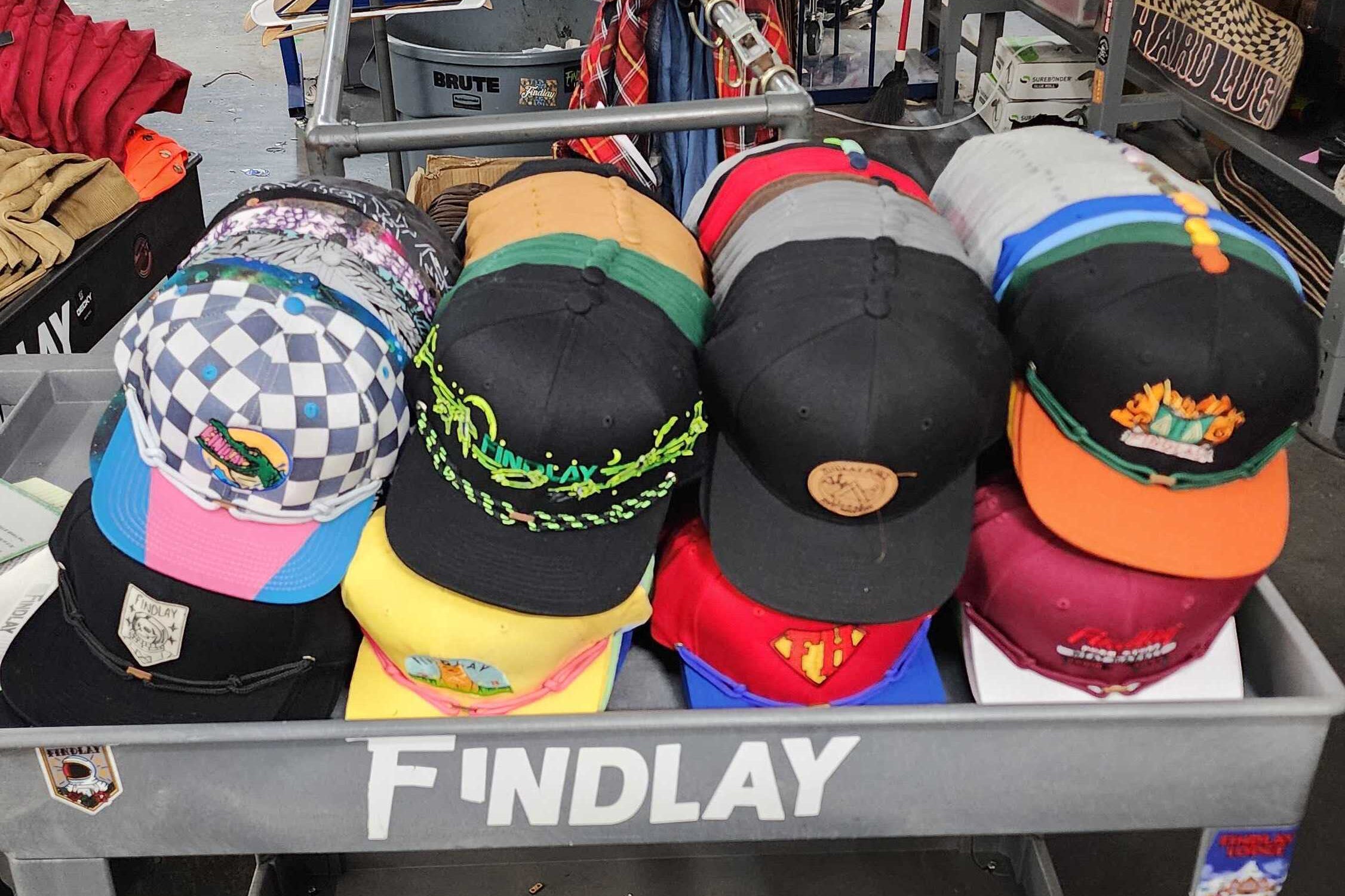 A collection of colorful baseball caps displayed on a metal shelf, with the brand name "FINDlay" written on the shelf.