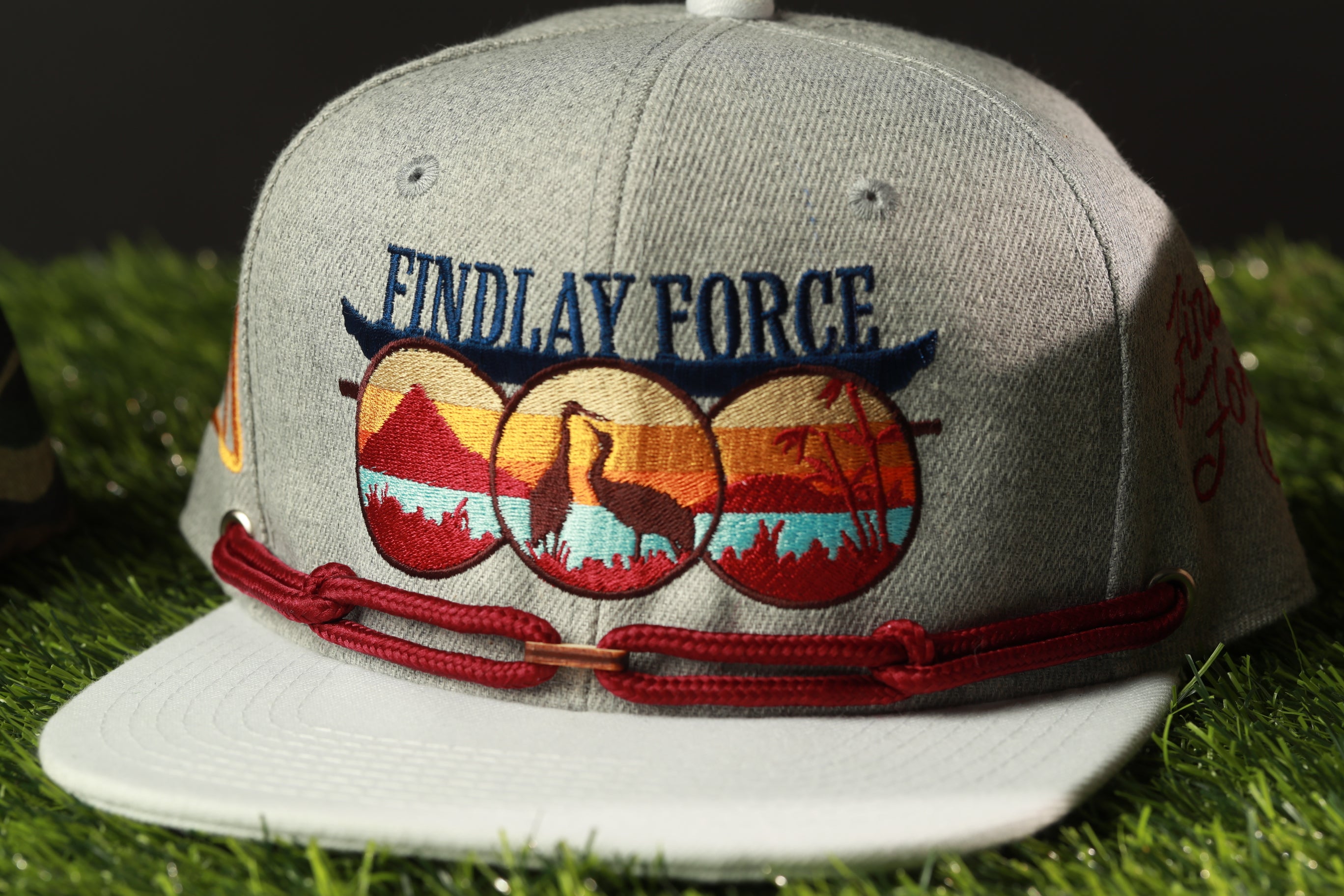 A gray baseball cap with a red band and a logo on the front, sitting on a grassy surface.