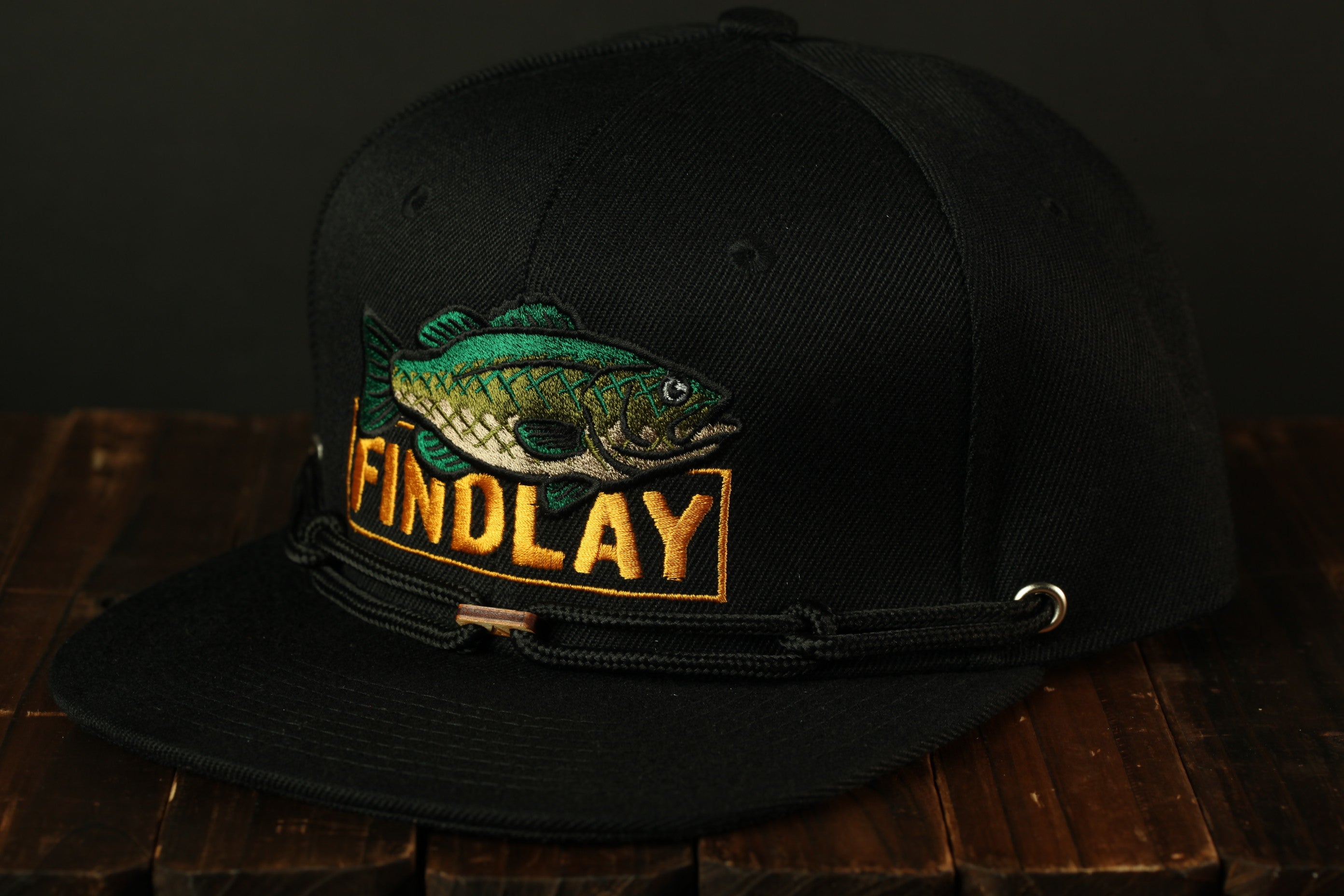A black baseball cap with a fish embroidered on it, sitting on a wooden surface.
