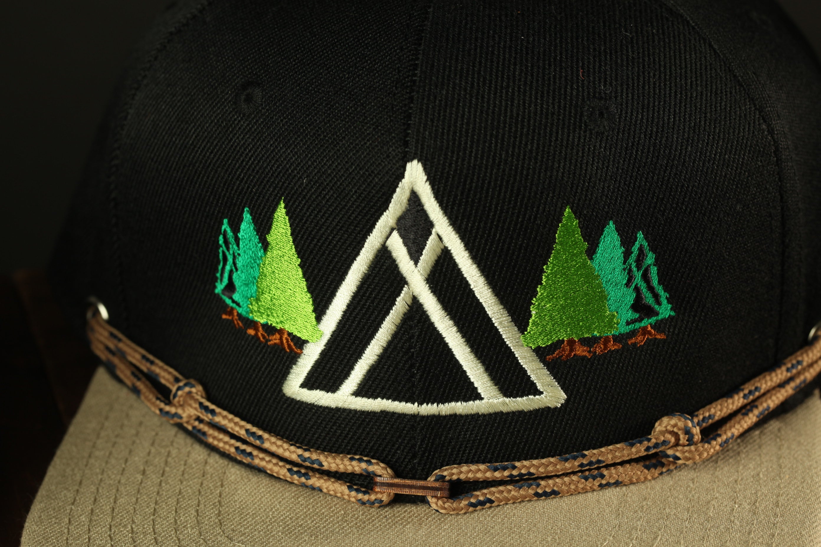 A close-up view of a black baseball cap with a white triangle design and green trees on either side.