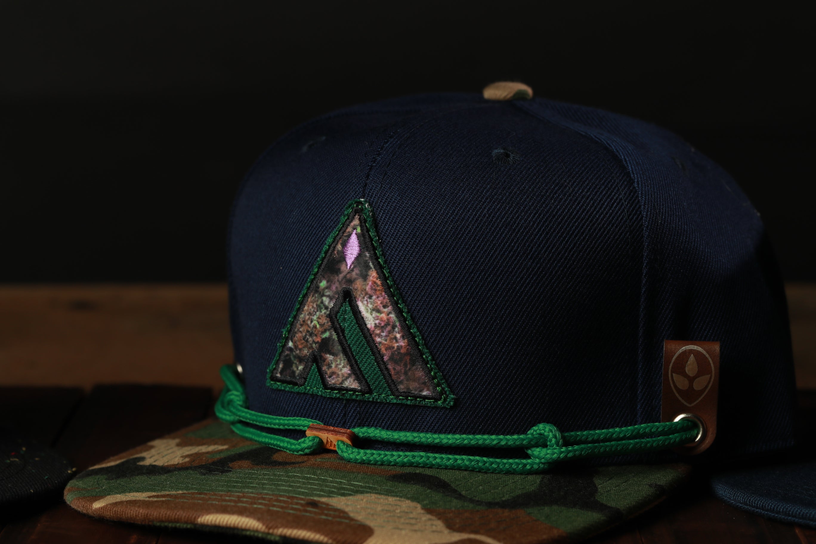 A navy blue baseball cap with a camouflage pattern and a triangular logo on the front, resting on a wooden surface.