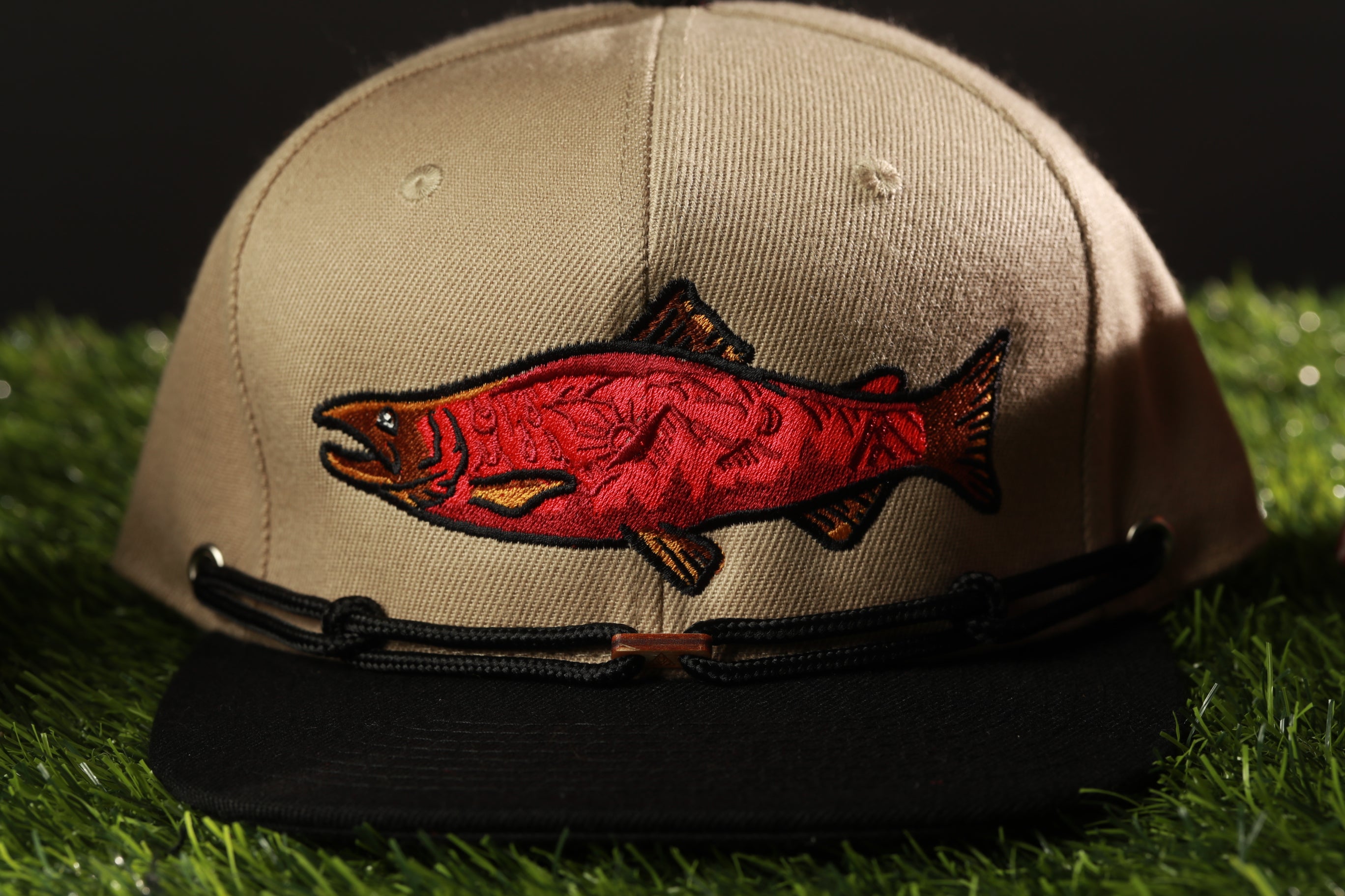 A beige baseball cap with a red fish embroidered on it, placed on a grassy surface.
