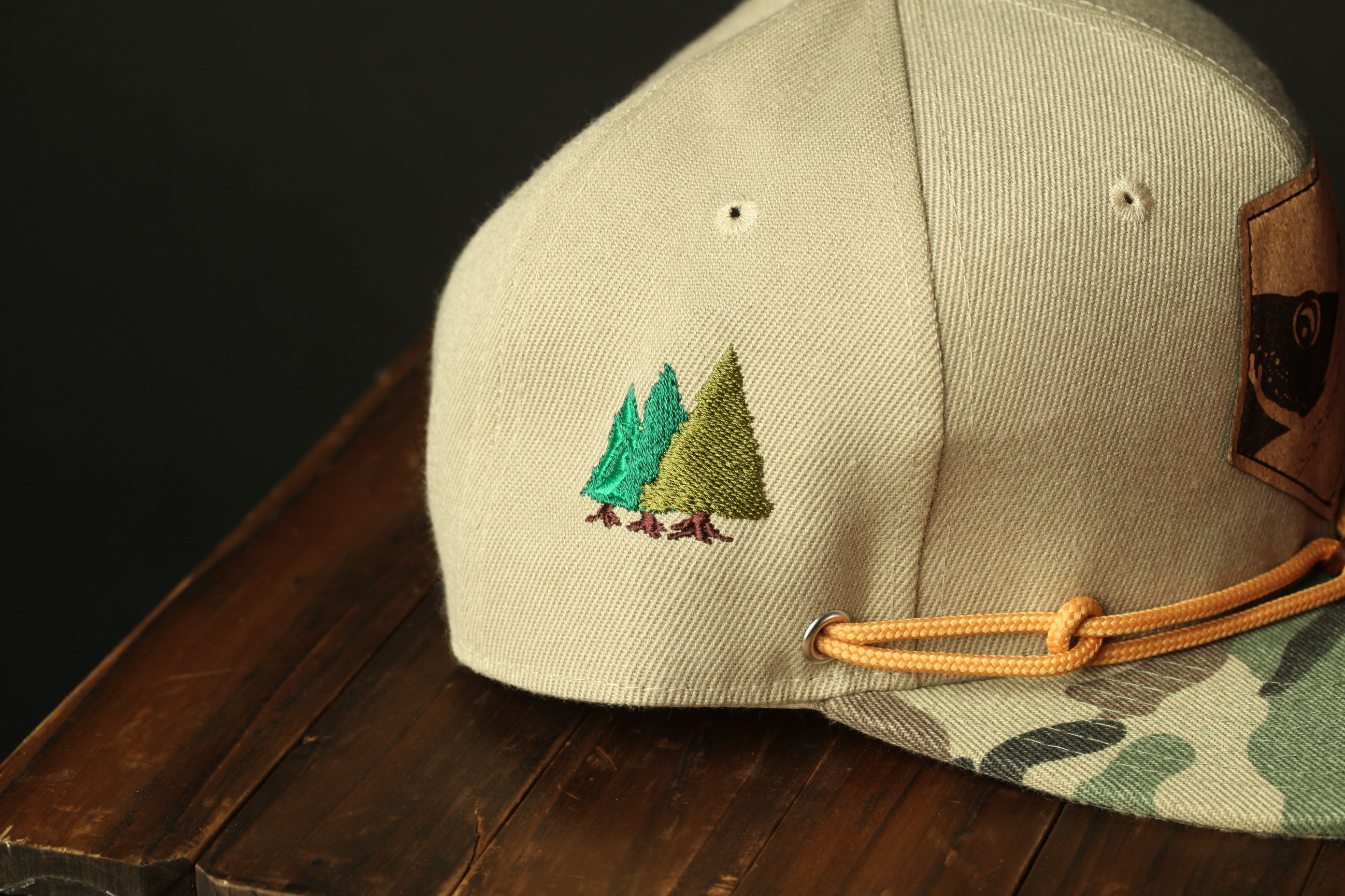 A beige baseball cap with a camouflage pattern and a small embroidered design of trees on the front.