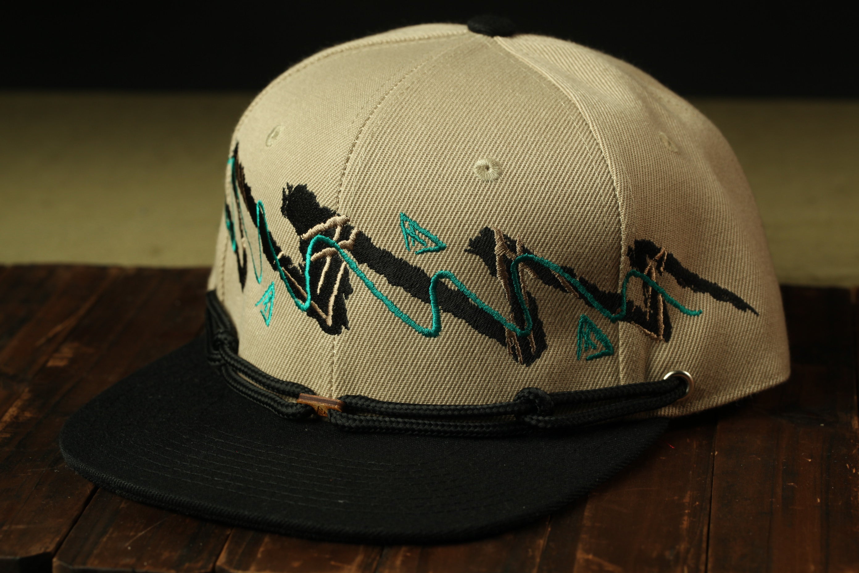 A beige baseball cap with a black brim and a graphic design on the front, sitting on a wooden surface.