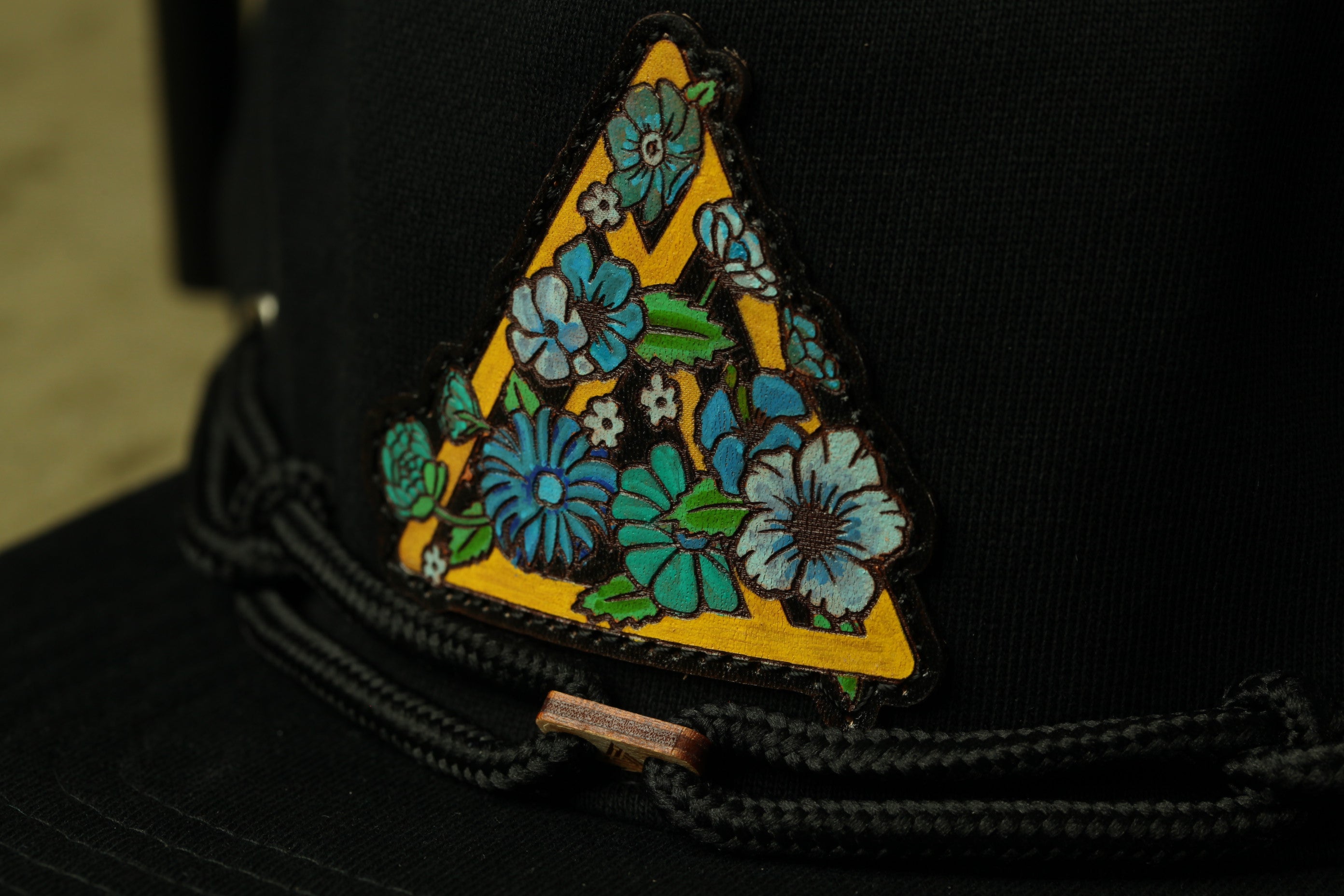 A close-up view of a black hat with a decorative triangular patch featuring a floral design in blue and green colors.