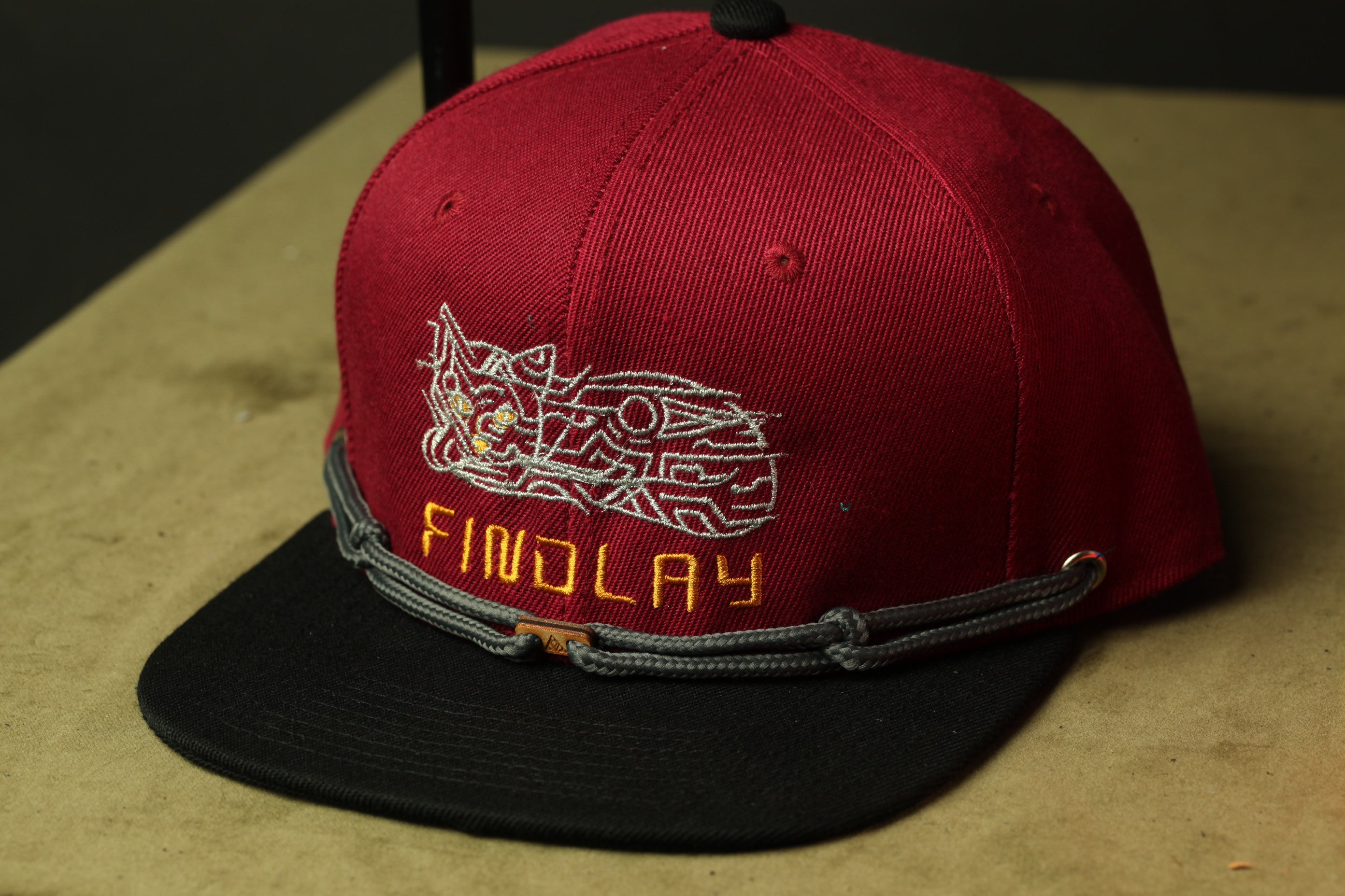 A red baseball cap with a black brim and a gray band, featuring a white embroidered design and the word "FINDOLAY" printed on it.