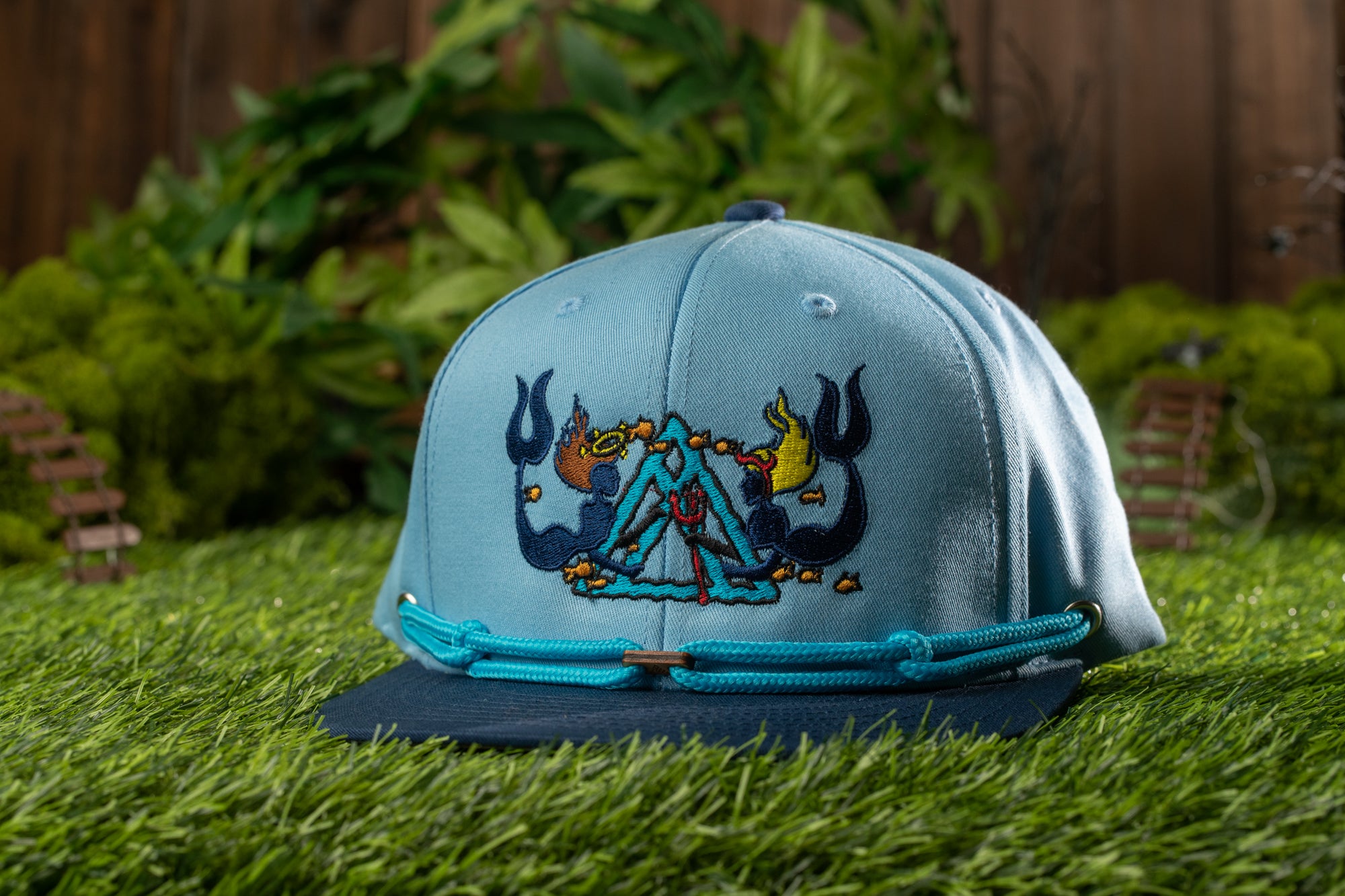 A blue baseball cap with a colorful, embroidered design sits on a grassy surface, with a wooden fence and greenery in the background.