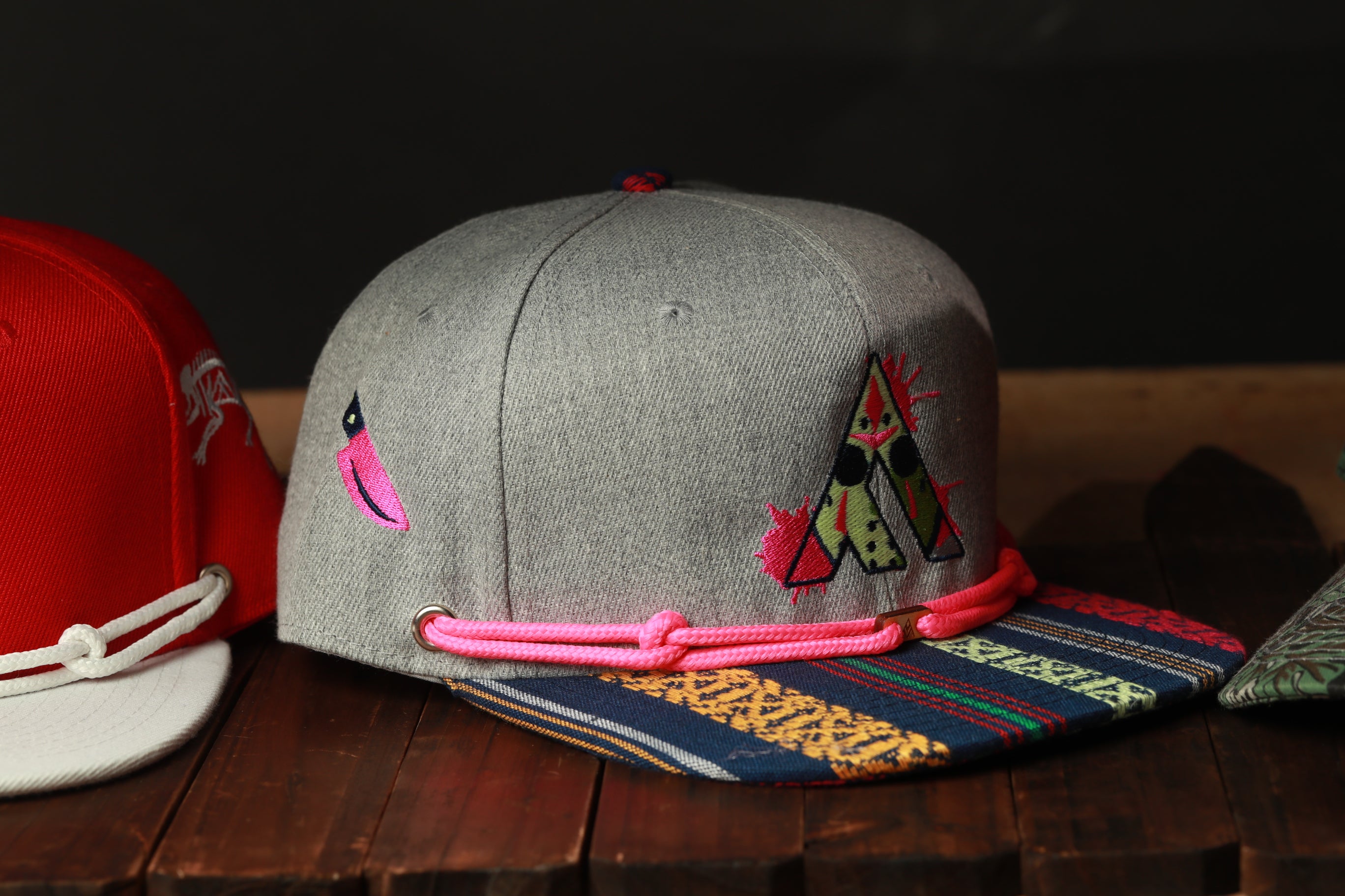 A gray baseball cap with colorful embroidered designs and a pink string is displayed on a wooden surface.