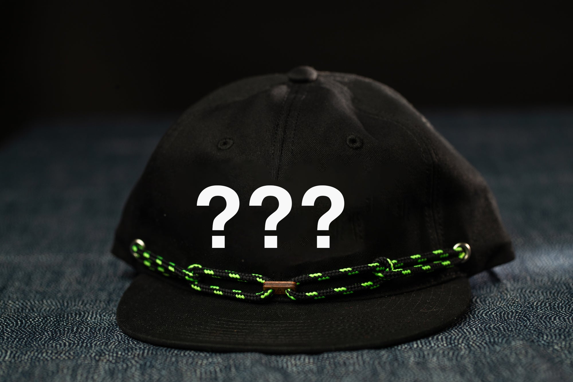 A black baseball cap with a green and black braided strap, featuring three question marks on the front.