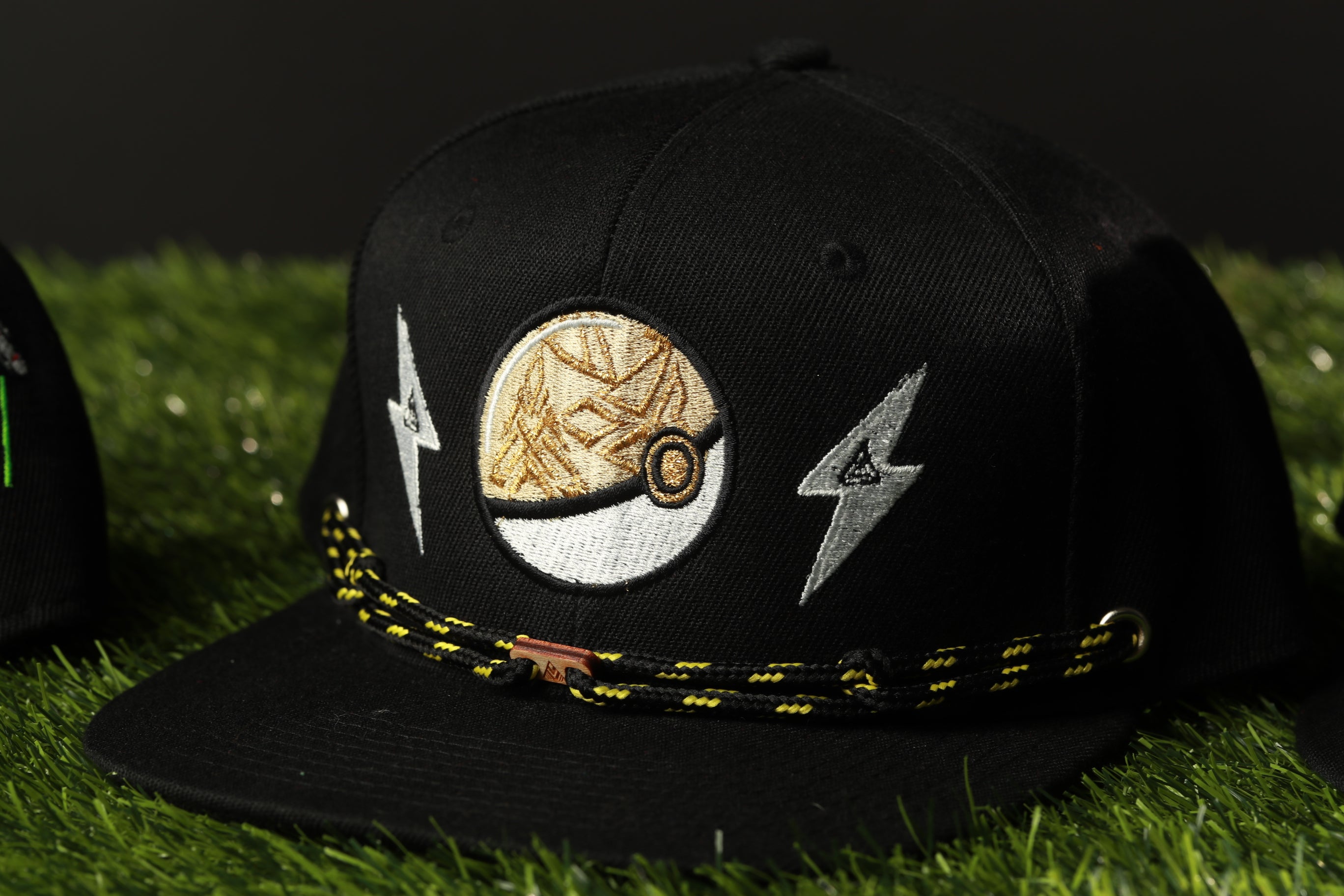 A black baseball cap with a gold and white embroidered design, featuring a lightning bolt symbol, is shown on a grassy surface.