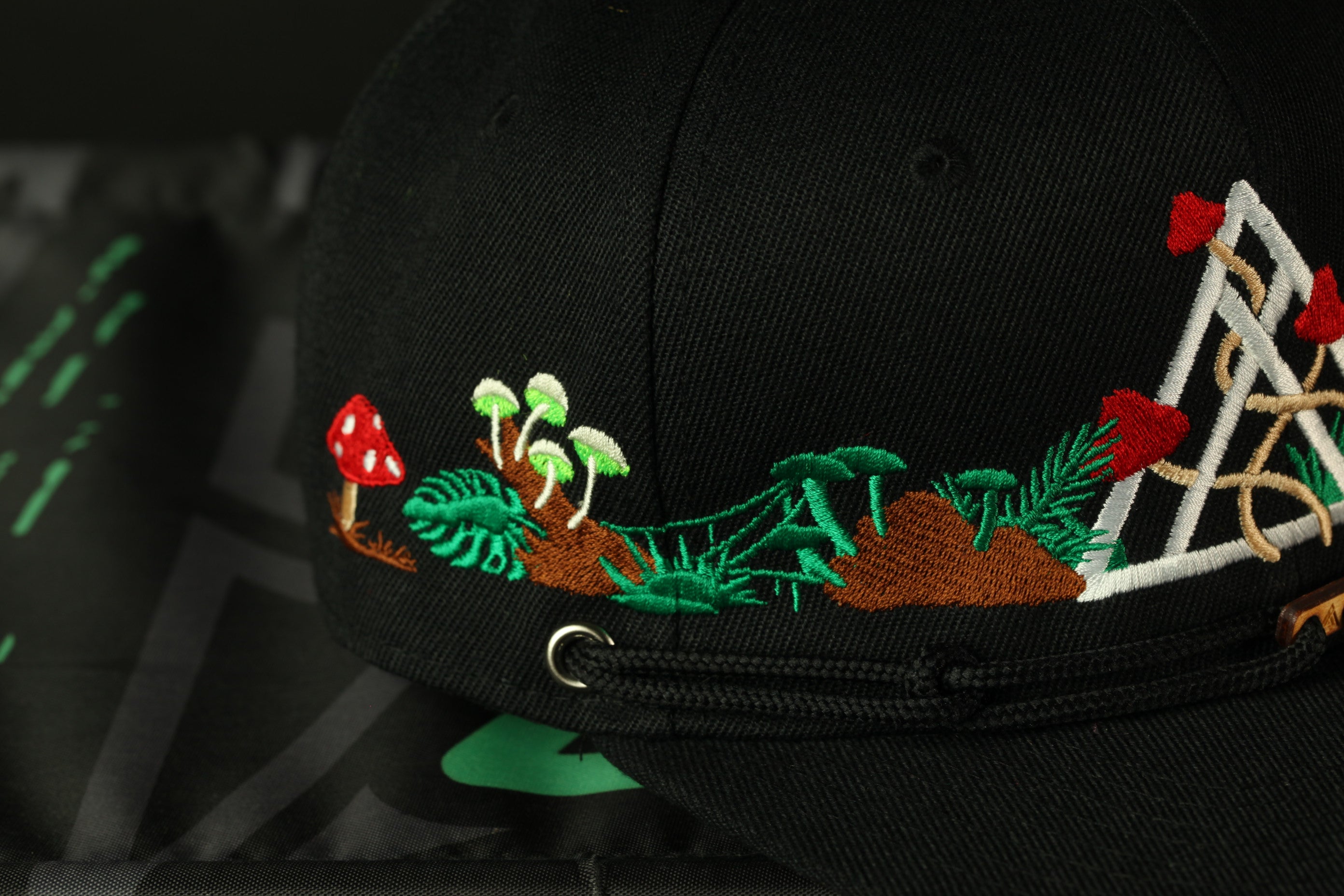 A close-up view of a black baseball cap with a detailed embroidered design featuring mushrooms, leaves, and other natural elements.